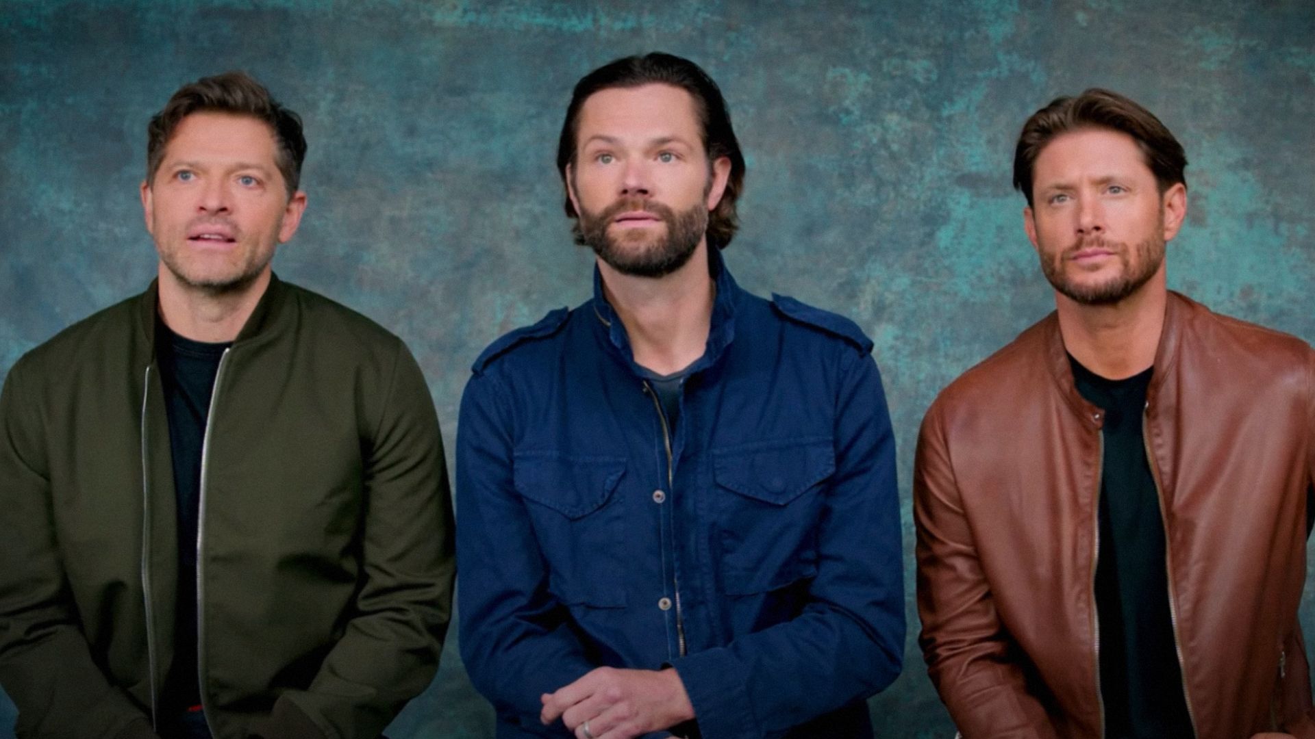 Actorii Misha Collins, Jared Padalecki și Jensen Ackles stau pe un fundal albastru purtând jachete colorate