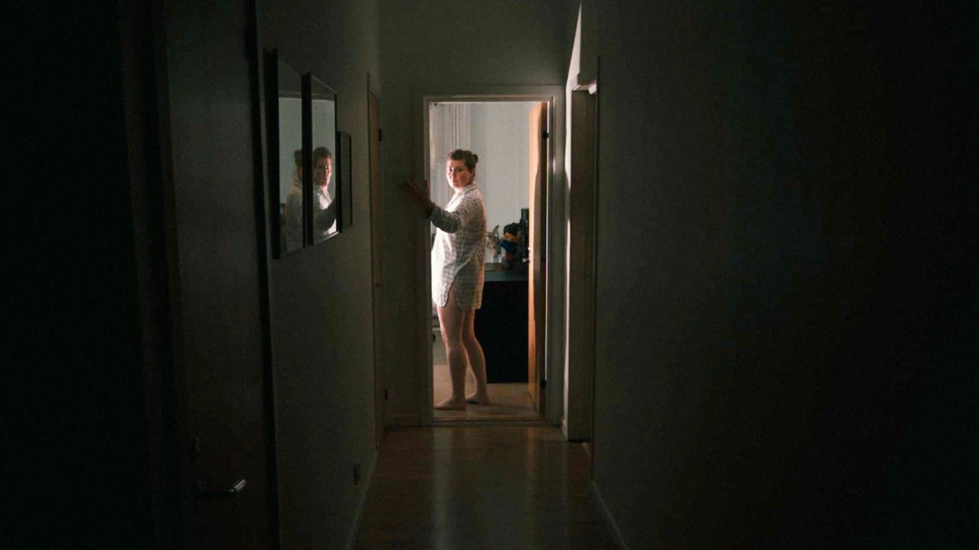 O femeie în cămașă de noapte stă în mijlocul holului apăsând întrerupătorul de lumină