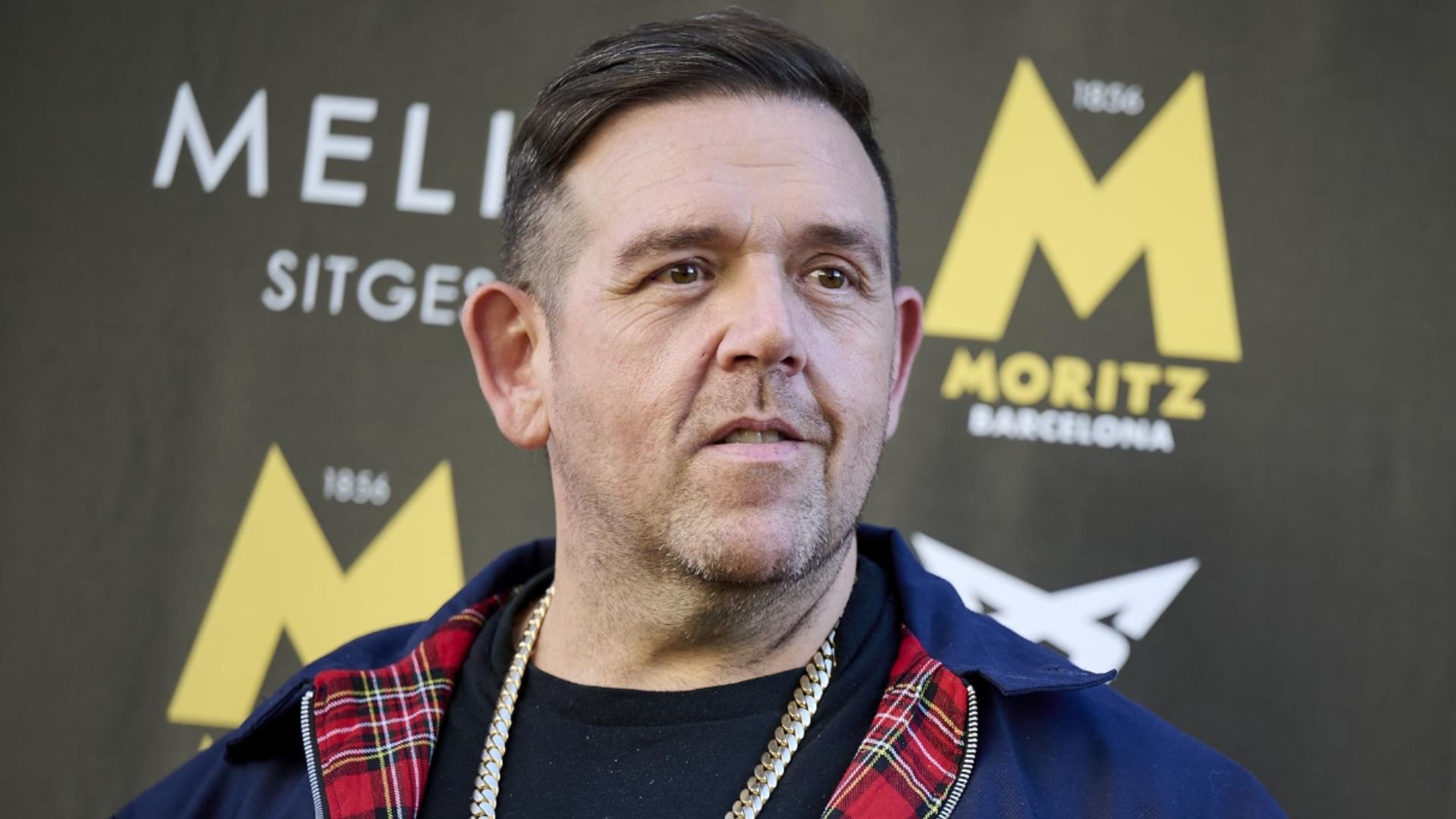 Actorul Nick Frost într-o jachetă albastră cu un lanț de aur în jurul gâtului zâmbește pe fondul unui display