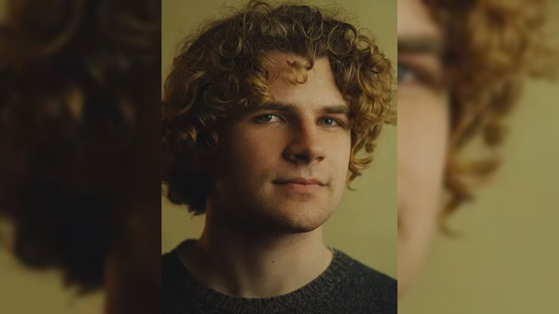 Un bărbat tânăr cu părul creț într-un pulover negru stă pe un fundal galben