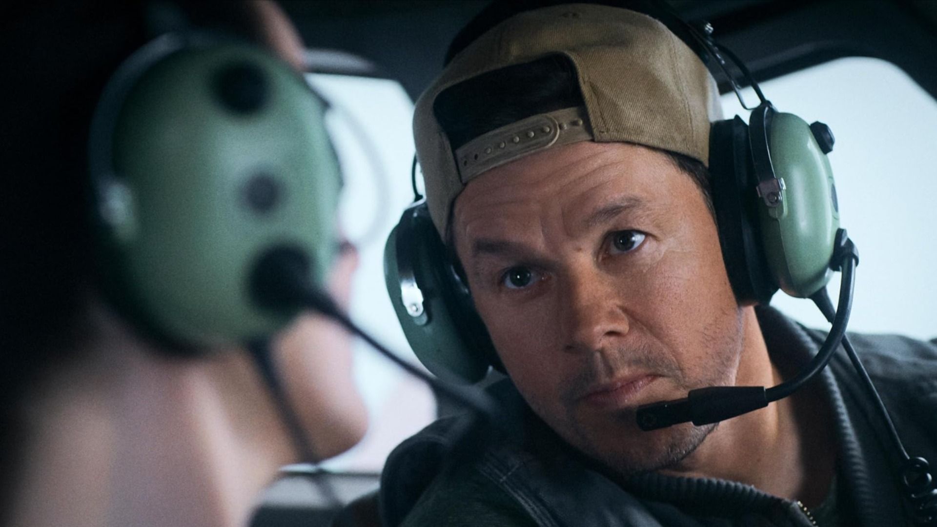 Actorul Mark Wahlberg purtând o șapcă și căști cu microfon stă într-un avion