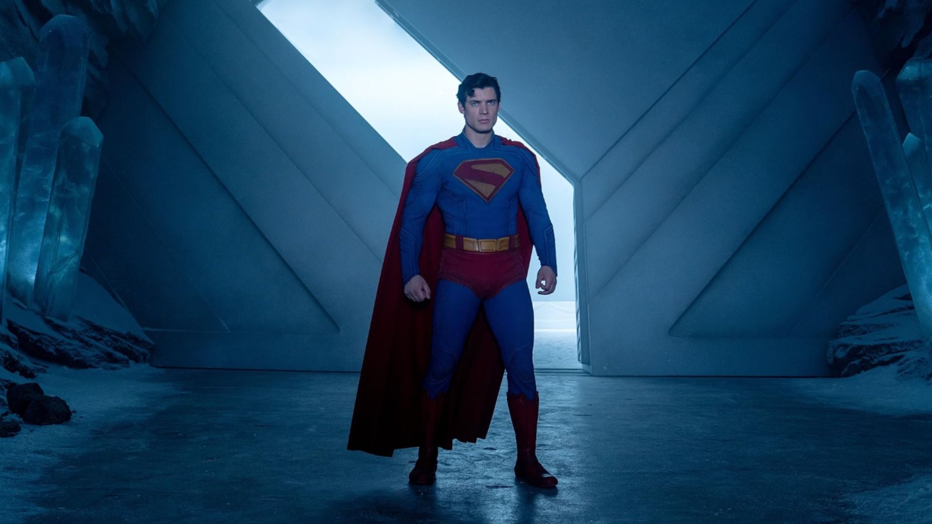 Actorul David Corenswet într-un costum de supererou albastru și roșu stă lângă niște porți mari