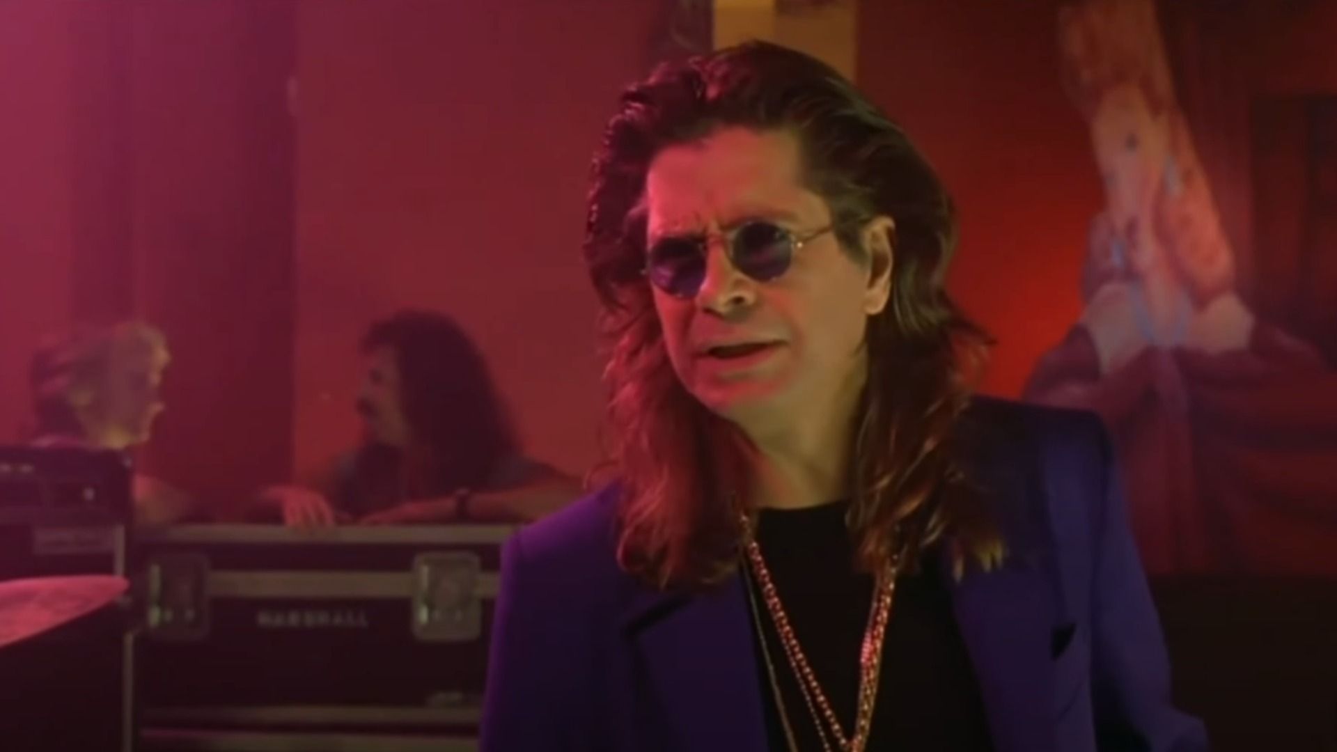 Cântărețul Ozzy Osbourne într-un blazer albastru și lanț de aur stă pe scenă