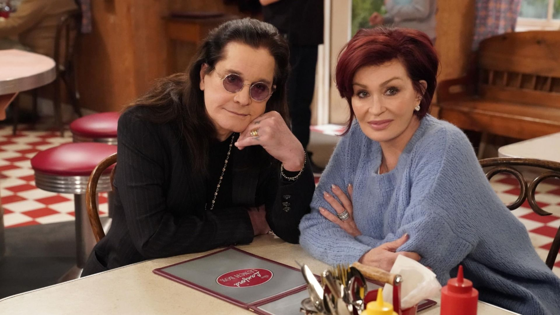 Cântărețul Ozzy Osbourne purtând ochelari și o jachetă neagră stă la masă cu soția sa