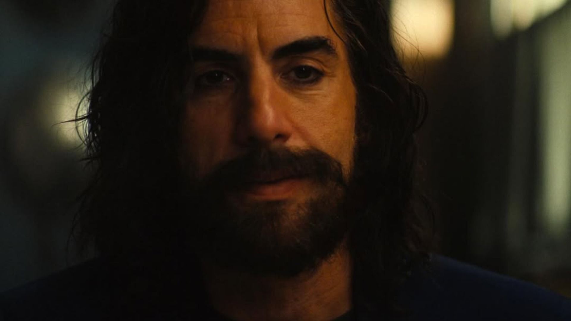 Actorul Sacha Baron Cohen cu barbă și păr lung stă pe un fundal estompat