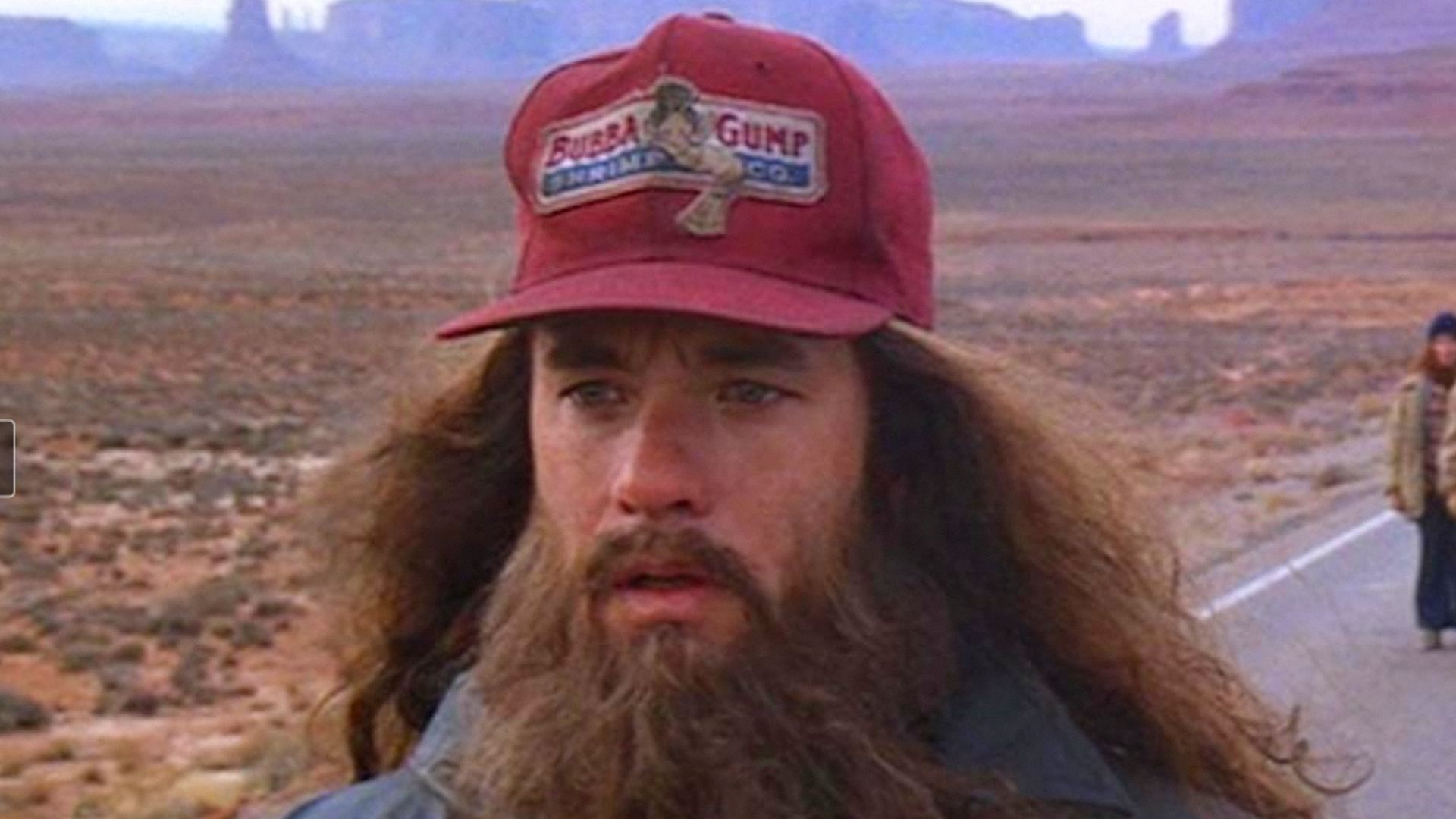 Actorul Tom Hanks cu părul lung și barbă stă purtând o șapcă roșie