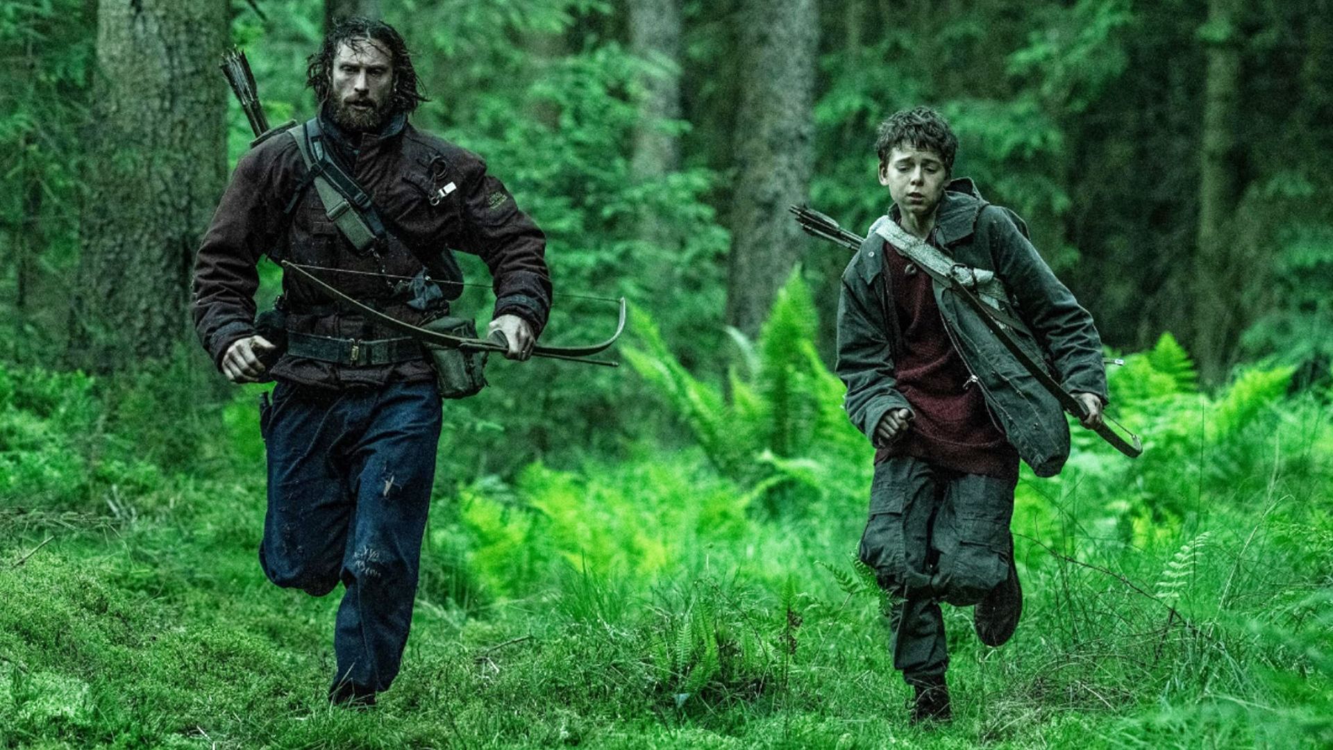 Actorul Aaron Taylor-Johnson în haine murdare aleargă prin pădure alături de un copil cu arc
