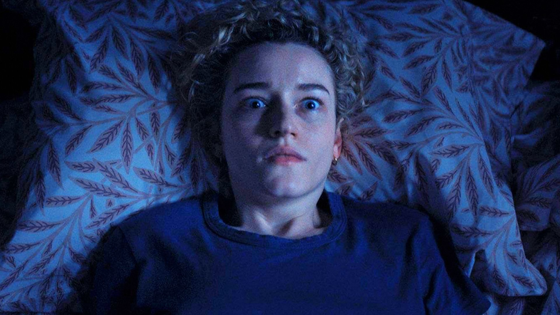 Actrița Julia Garner într-un pulover albastru stă pe pat, speriată de ceva