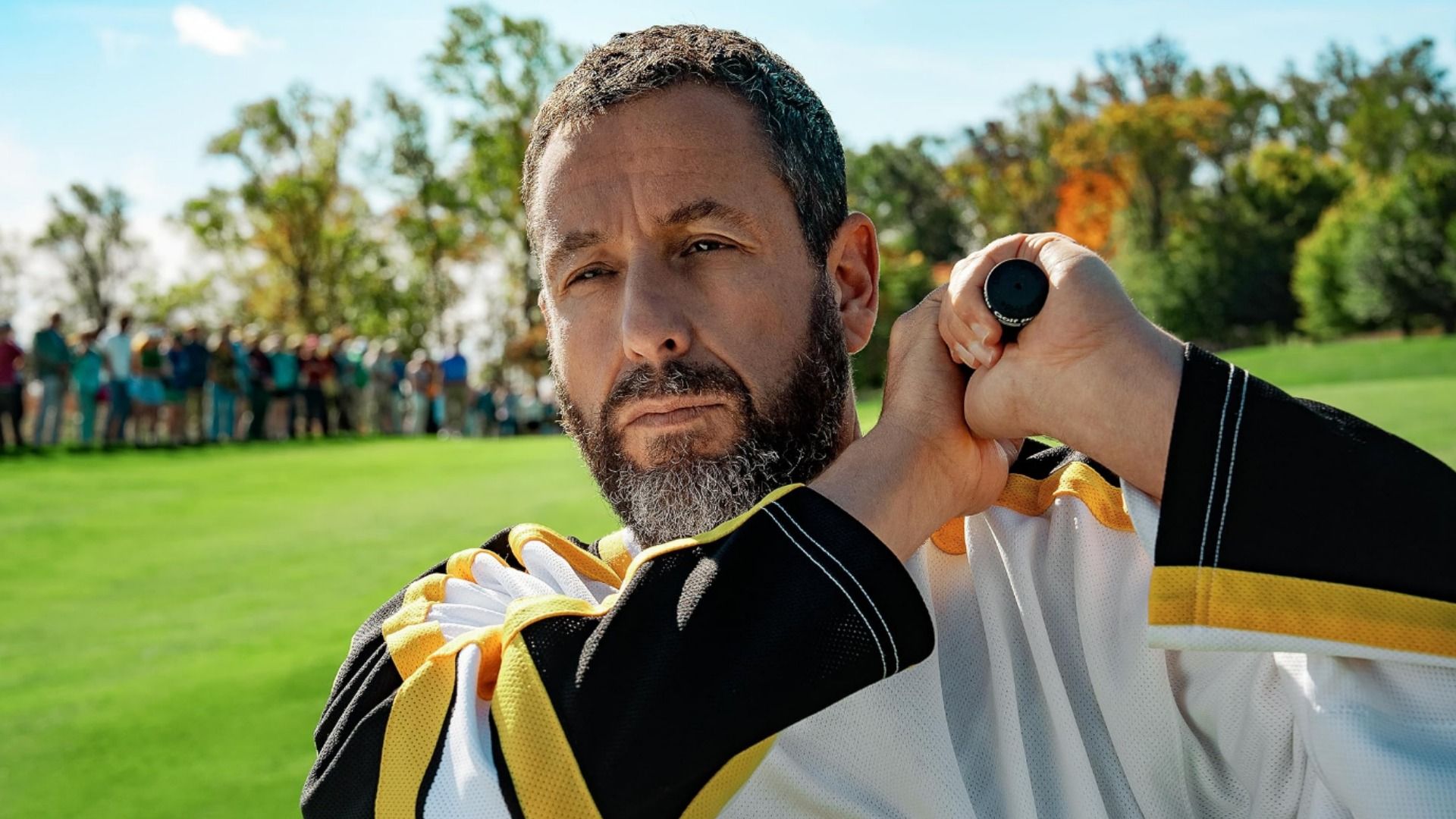 Actorul Adam Sandler ținând o crosă de golf pe un teren de golf