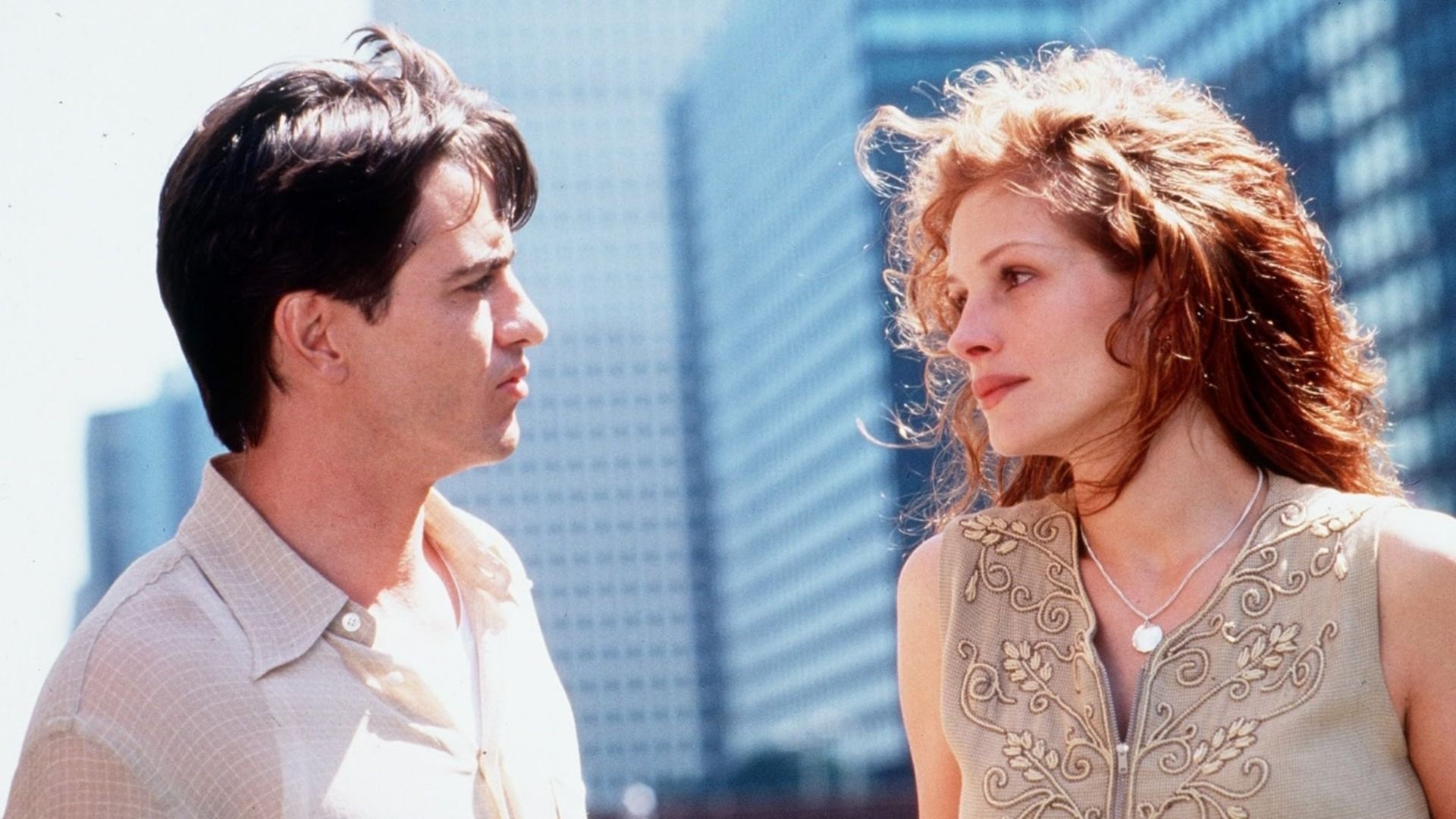 Actorul Dermot Mulroney într-o cămașă deschisă se uită la actrița Julia Roberts pe fundalul unor clădiri înalte