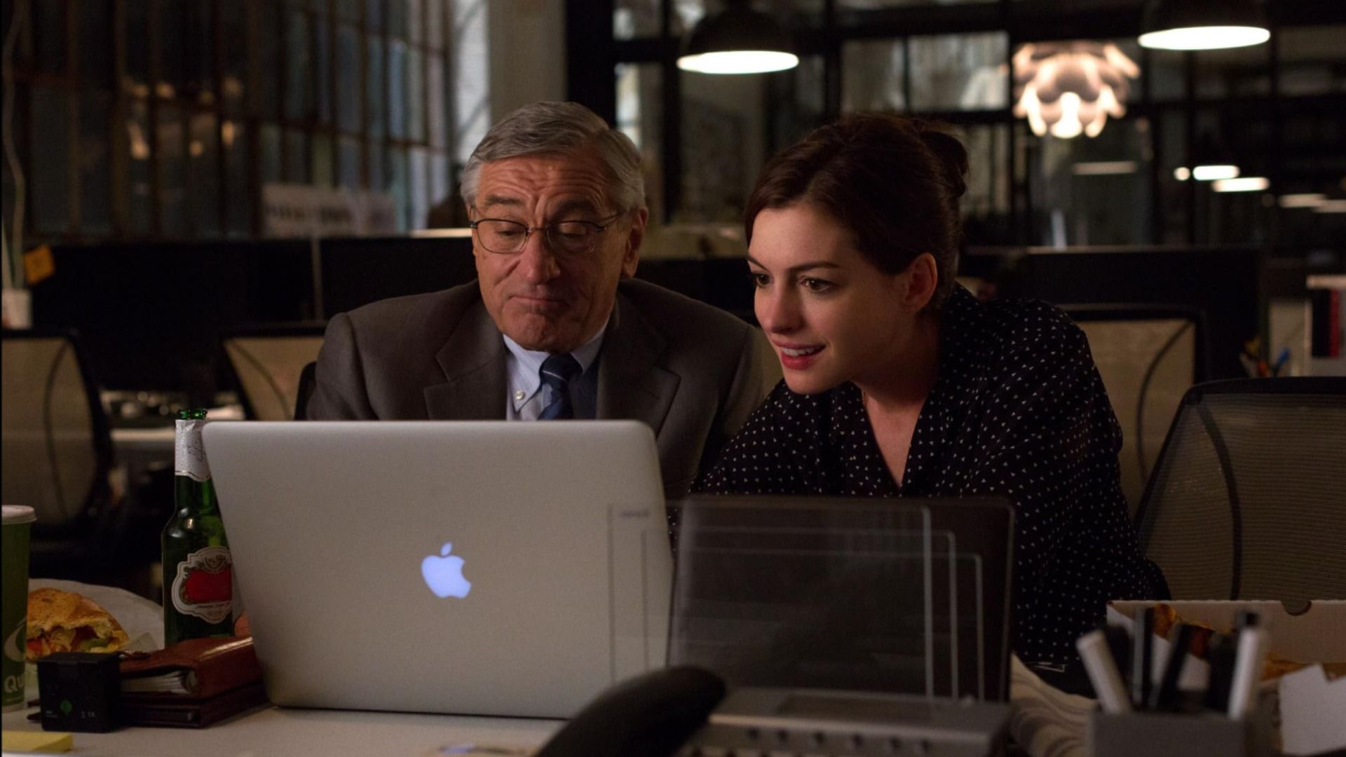 Actorul Robert De Niro într-un sacou gri și actrița Anne Hathaway într-o rochie neagră stând la masă