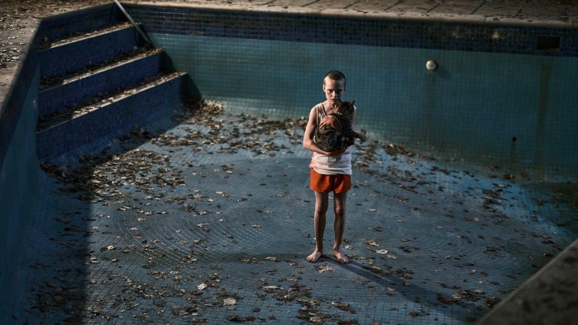 Un băiat în pantaloni scurți portocalii stă pe fundul unei piscine ținând o pisică în brațe
