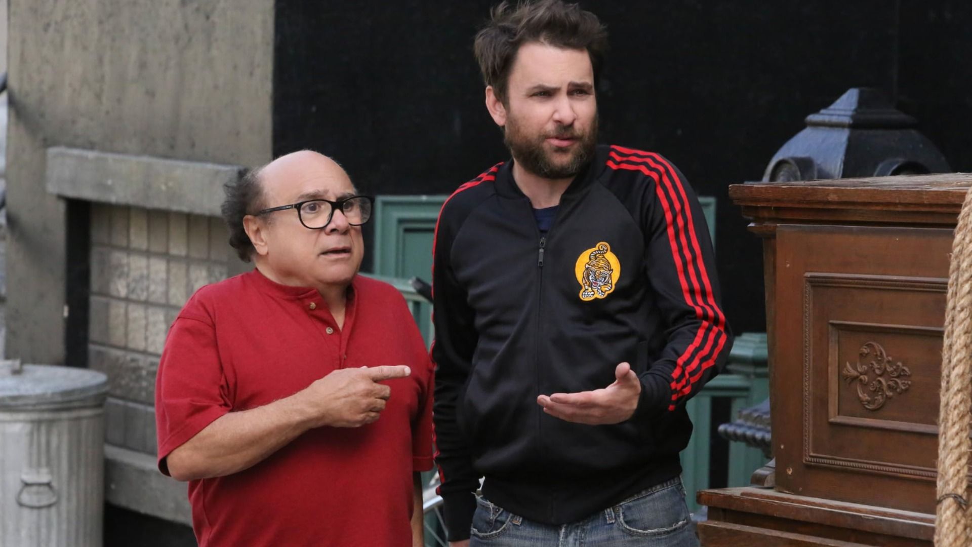 Actorul Danny DeVito într-o cămașă roșie îi arată cu degetul unui bărbat în echipament sportiv