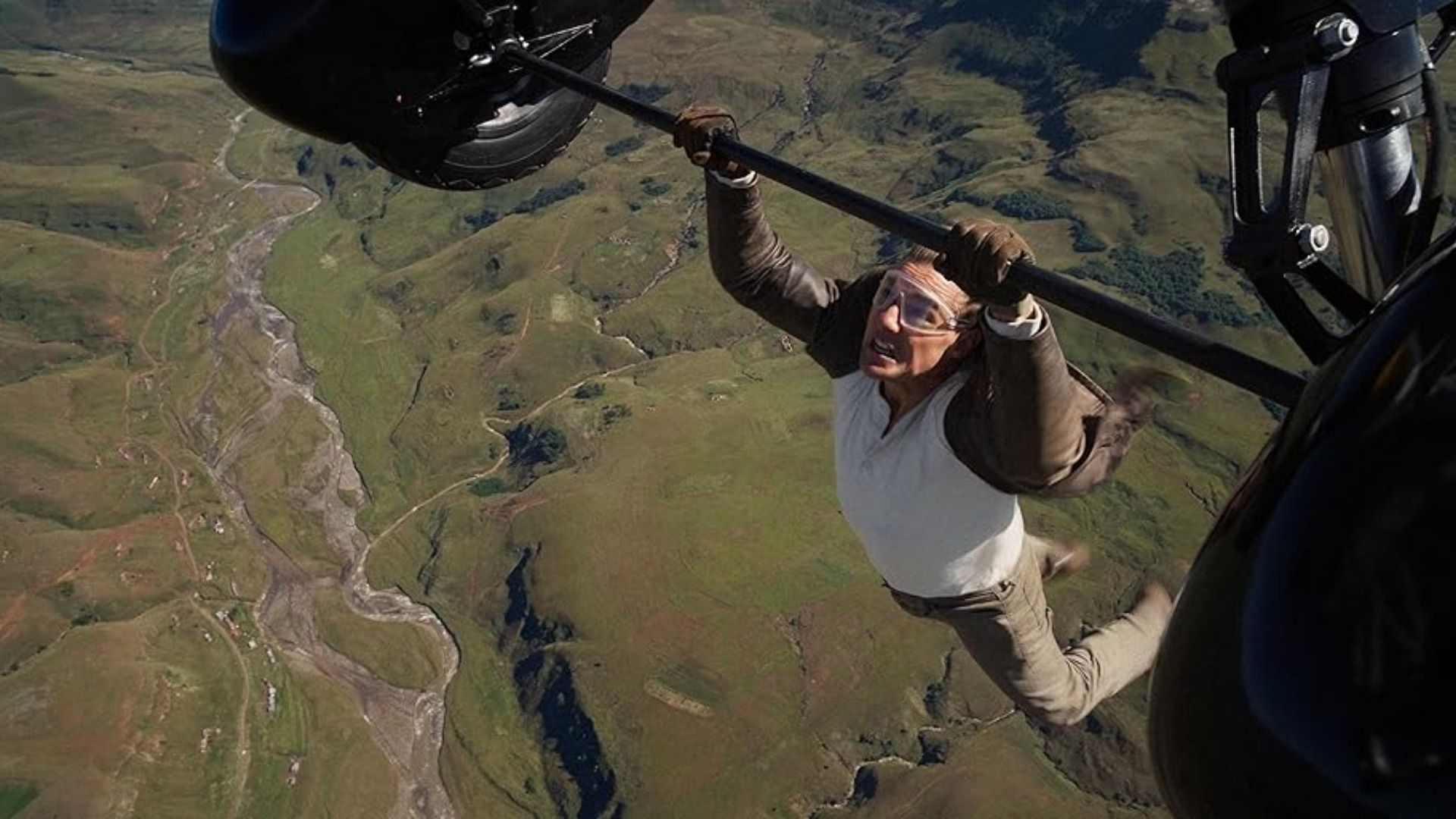 Actorul Tom Cruise în jachetă maro se agață de roțile unui avion în zbor