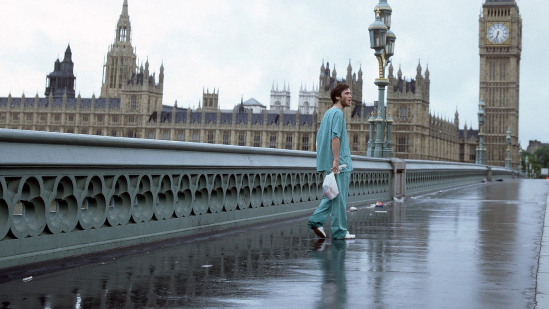 Actorul Cillian Murphy în haine albastre stă pe un pod gol cu turnul cu ceas Big Ben din Londra vizibil în apropiere