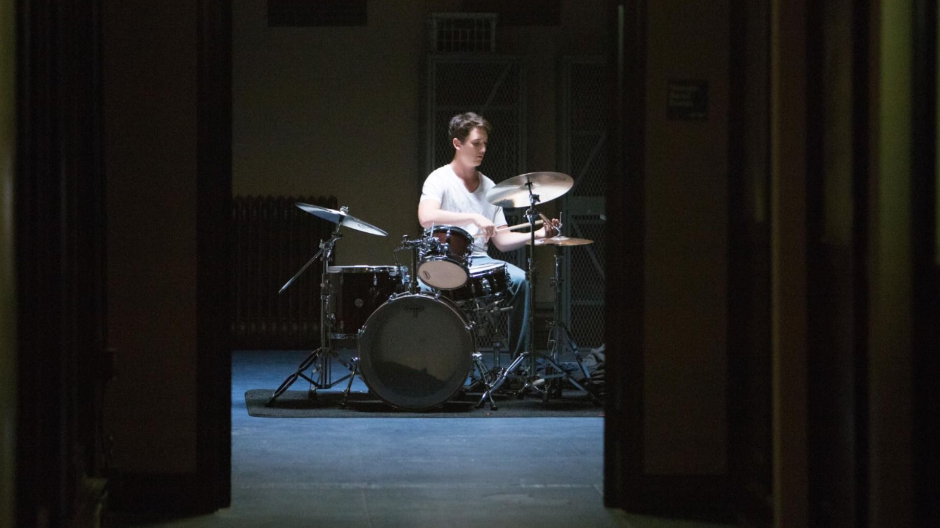 Lumină estompată, actorul Miles Teller cântă la tobe într-un tricou alb