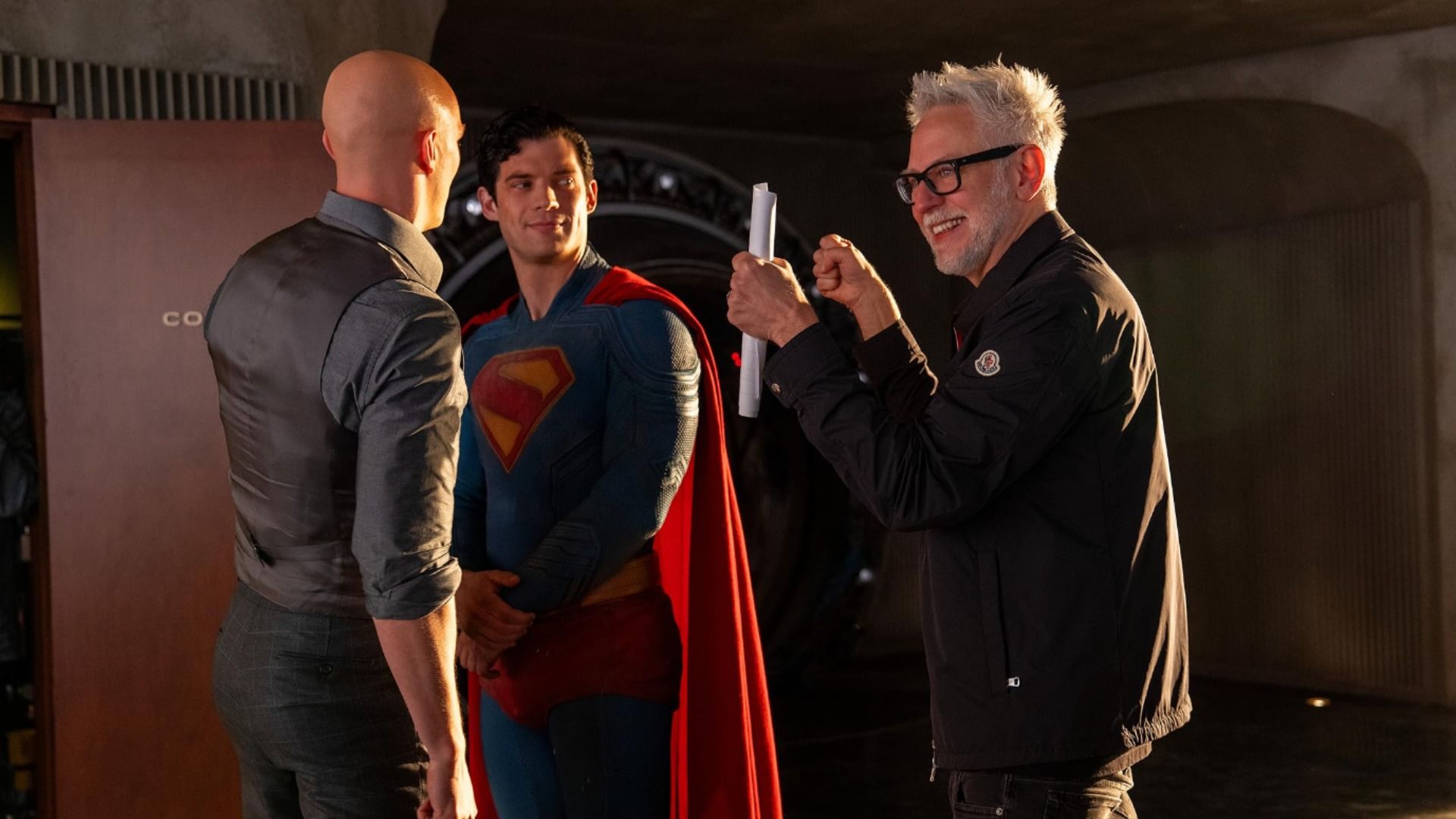 Regizorul James Gunn ținând hârtii vorbește cu actorul David Corenswet în costum de Superman și cu Nicholas Hoult care este chel