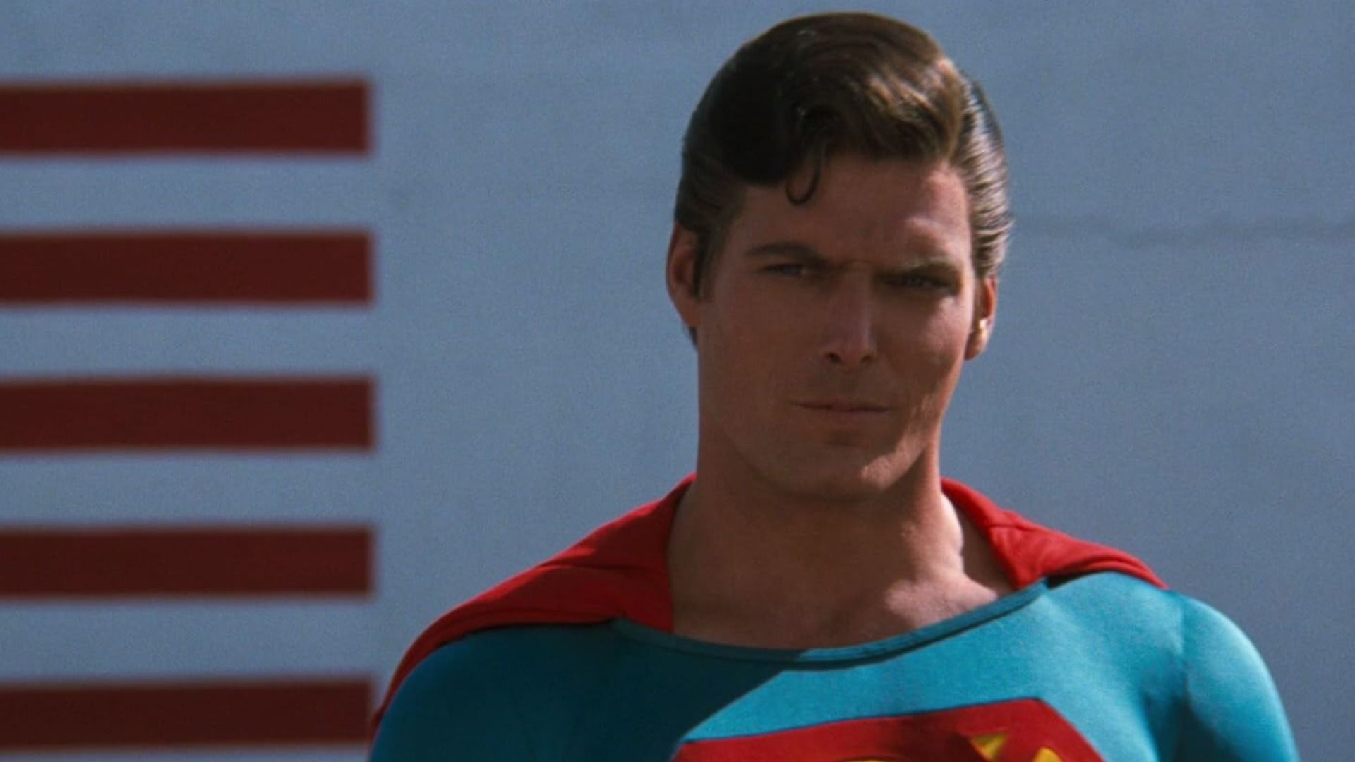 Actorul Christopher Reeve în costumul albastru și roșu al lui Superman se uită amenințător într-o parte