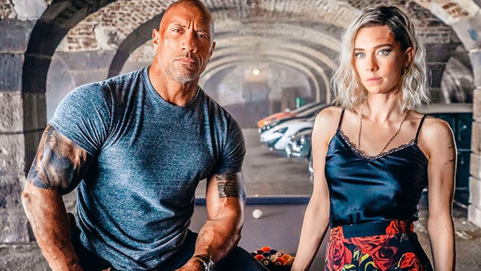 Dwayne Johnson într-un tricou strâmt și Vanessa Kirby într-un maiou și fustă cu model pozează pe un fundal cu arcuri și mașini sport