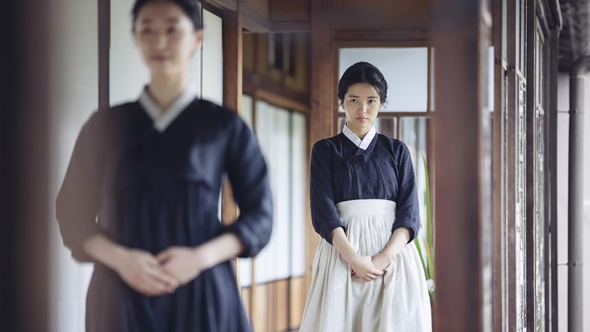 O fată în hanbok negru cu șorț alb stă în coridor, privind înainte cu o expresie serioasă