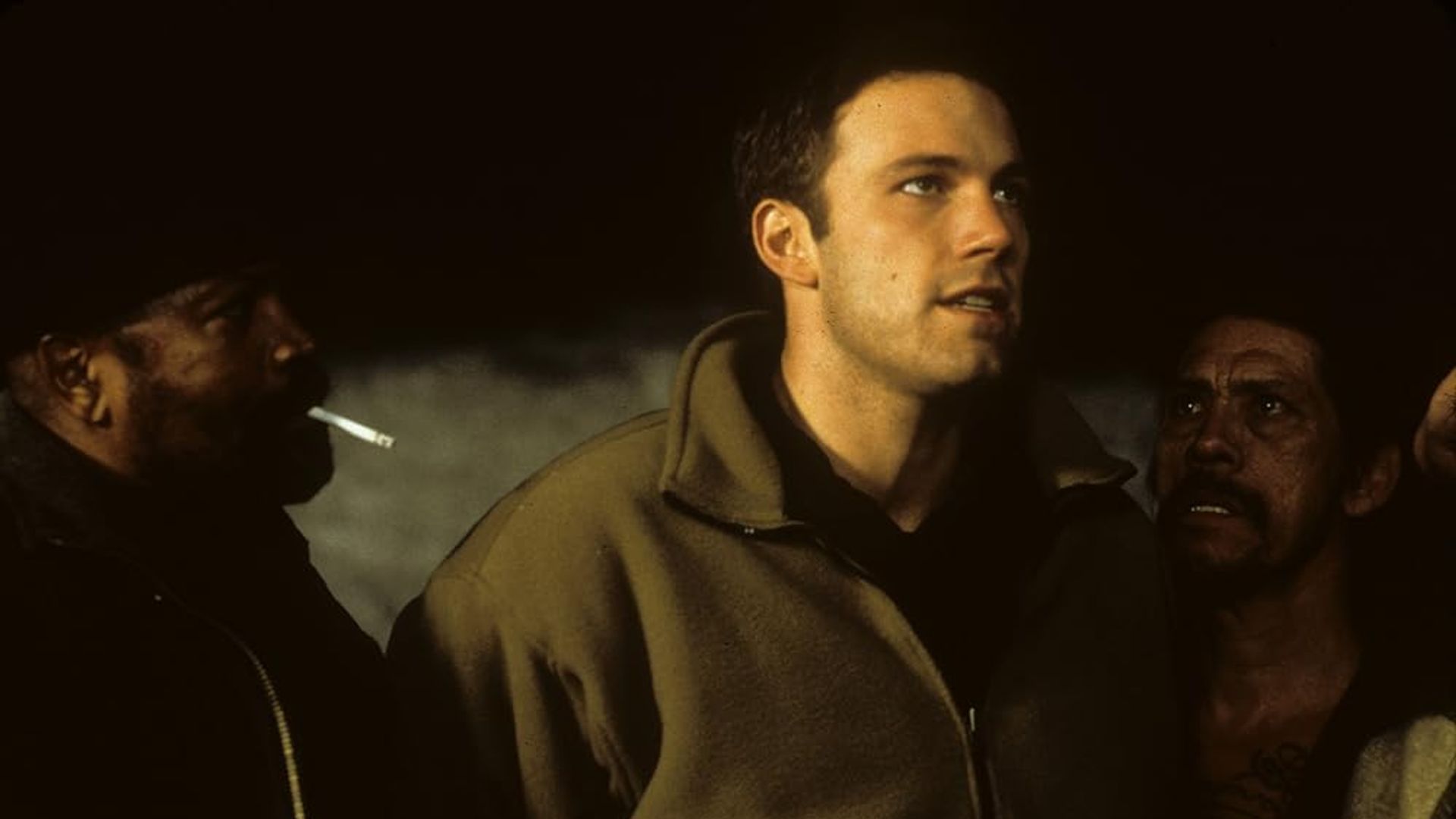 Ben Affleck într-o jachetă bej stă între două persoane, una dintre ele fumând, într-un cadru slab luminat