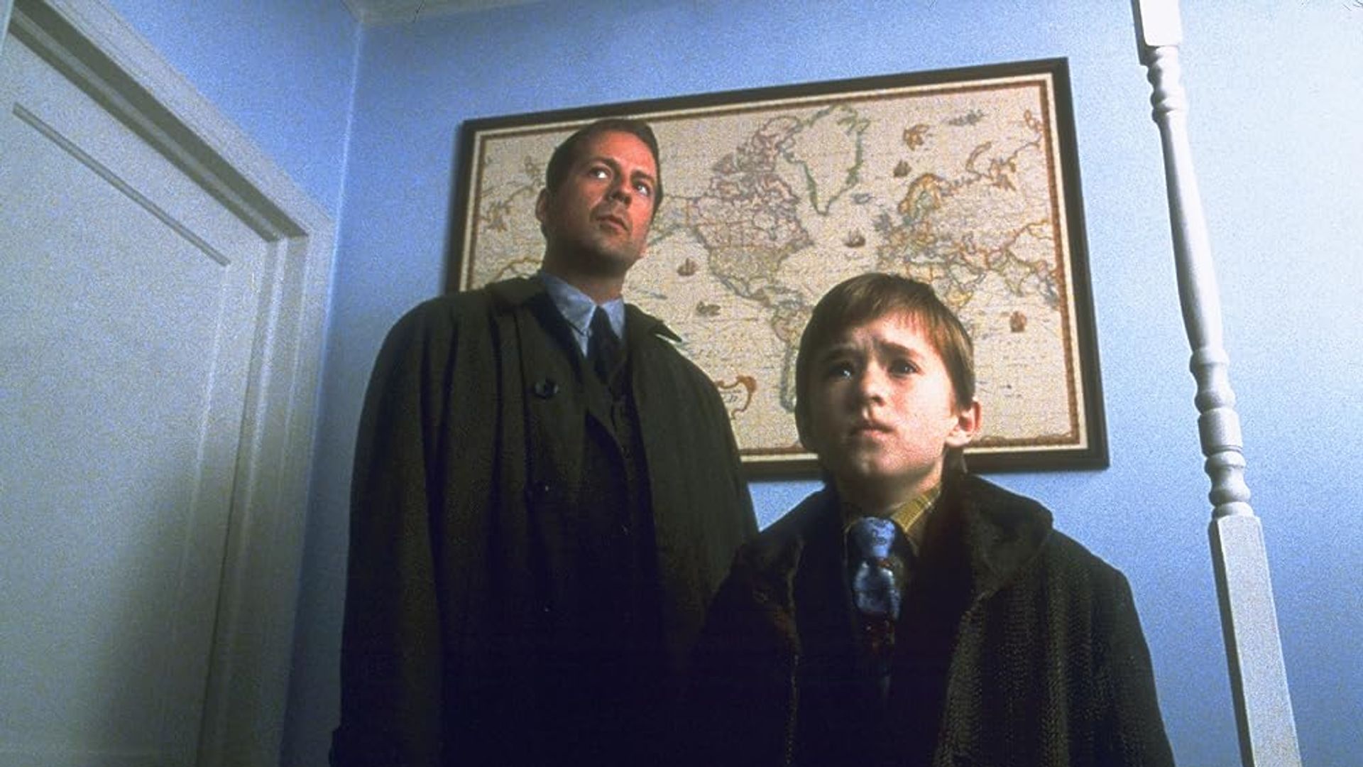 Bruce Willis și Haley Joel Osment stând într-un coridor cu o hartă a lumii agățată pe perete în spatele lor
