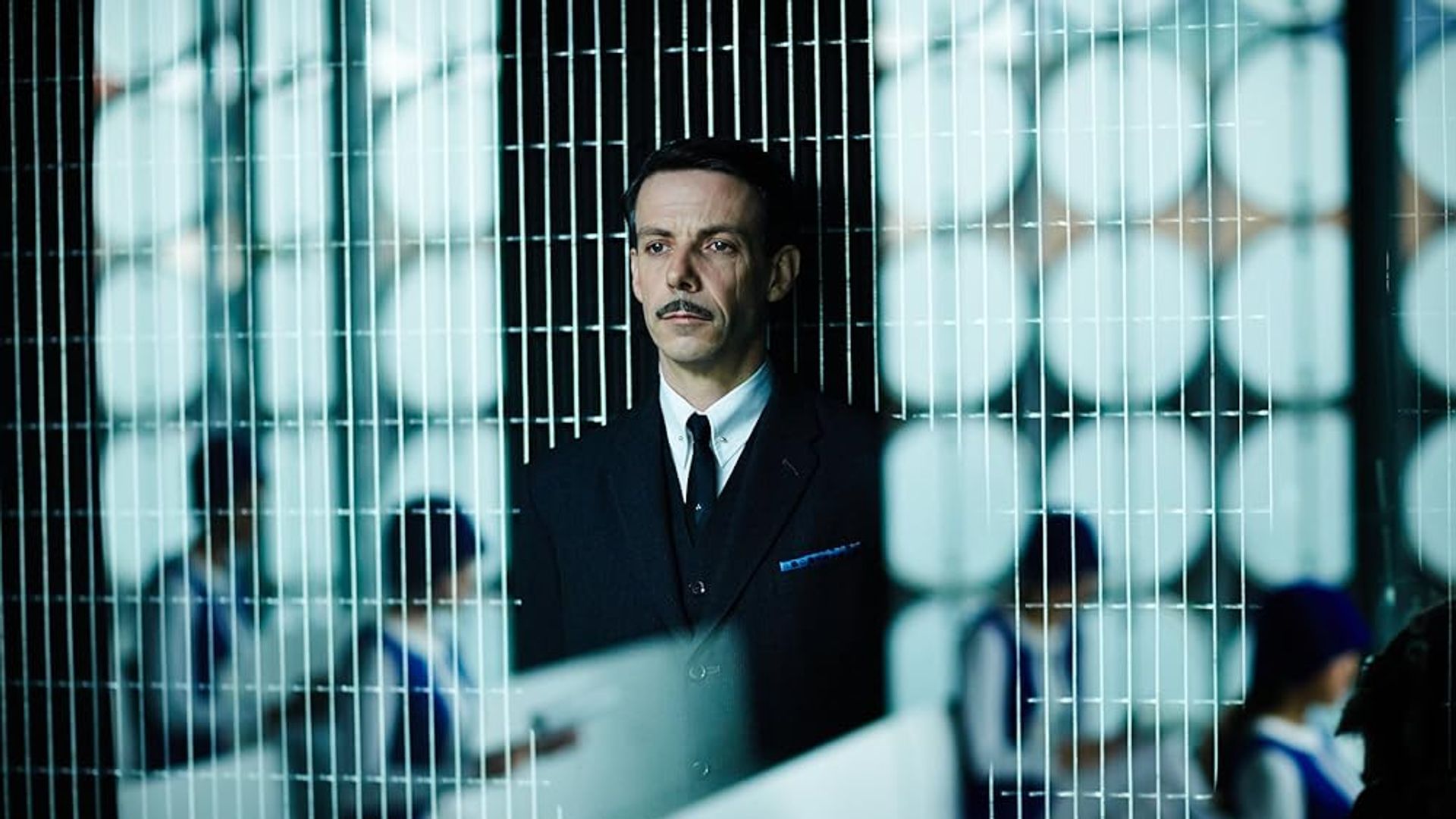 Ethan Hawke în costum cu mustață stând în spatele unei partiții din plasă cu oameni în uniformă lucrând la birouri în fundal