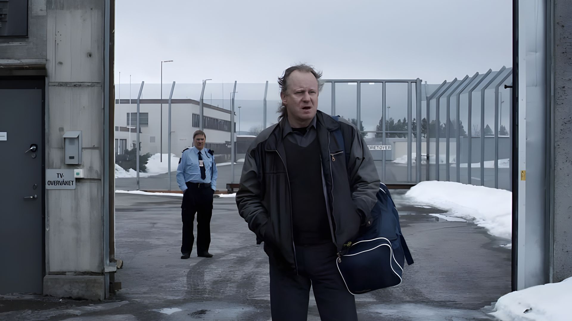 Stellan Skarsgård purtând o geantă iese prin porțile închisorii, cu un gardian în fundal, clădiri și zăpadă vizibile dincolo de gard