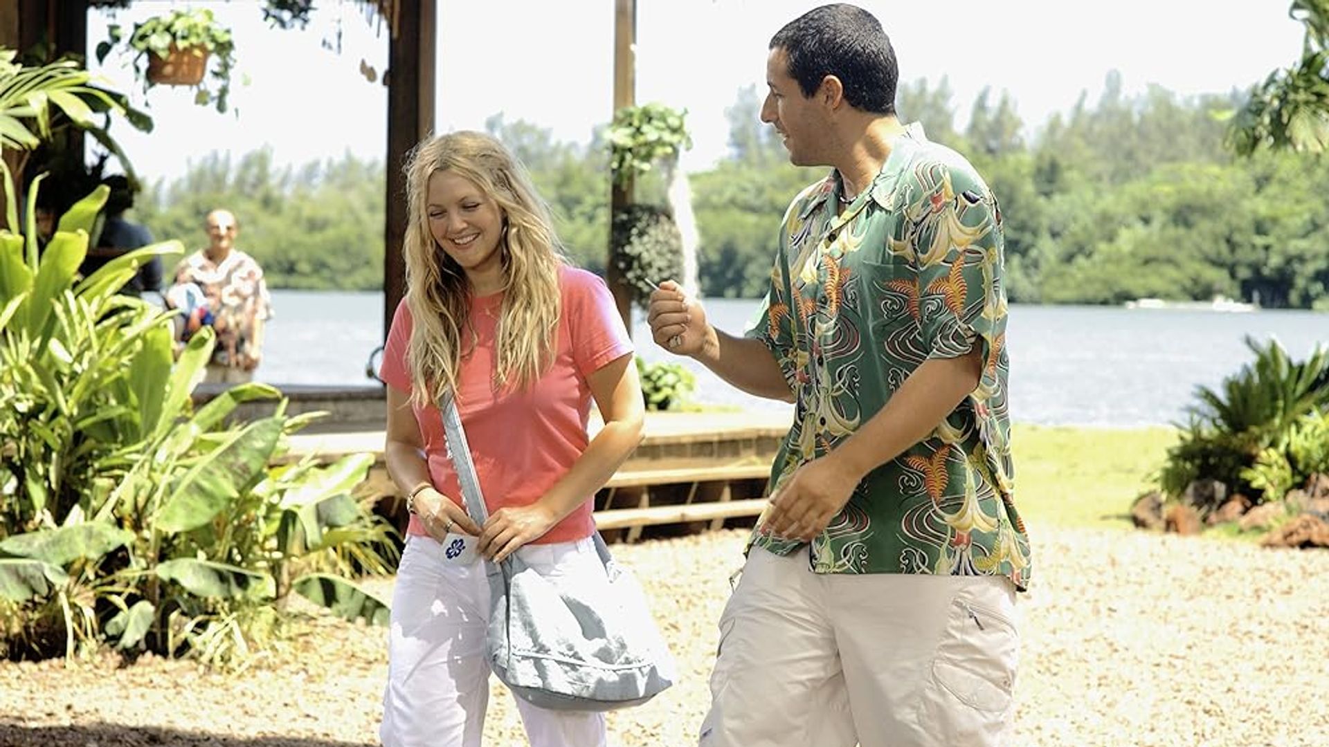Drew Barrymore și Adam Sandler vorbind pe o plajă însorită cu verdeață și apă în fundal