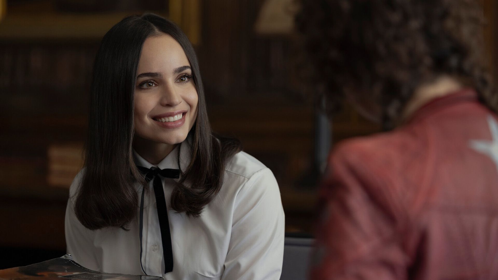 Sofia Carson într-o bluză albă cu o fundiță neagră la gât zâmbind larg