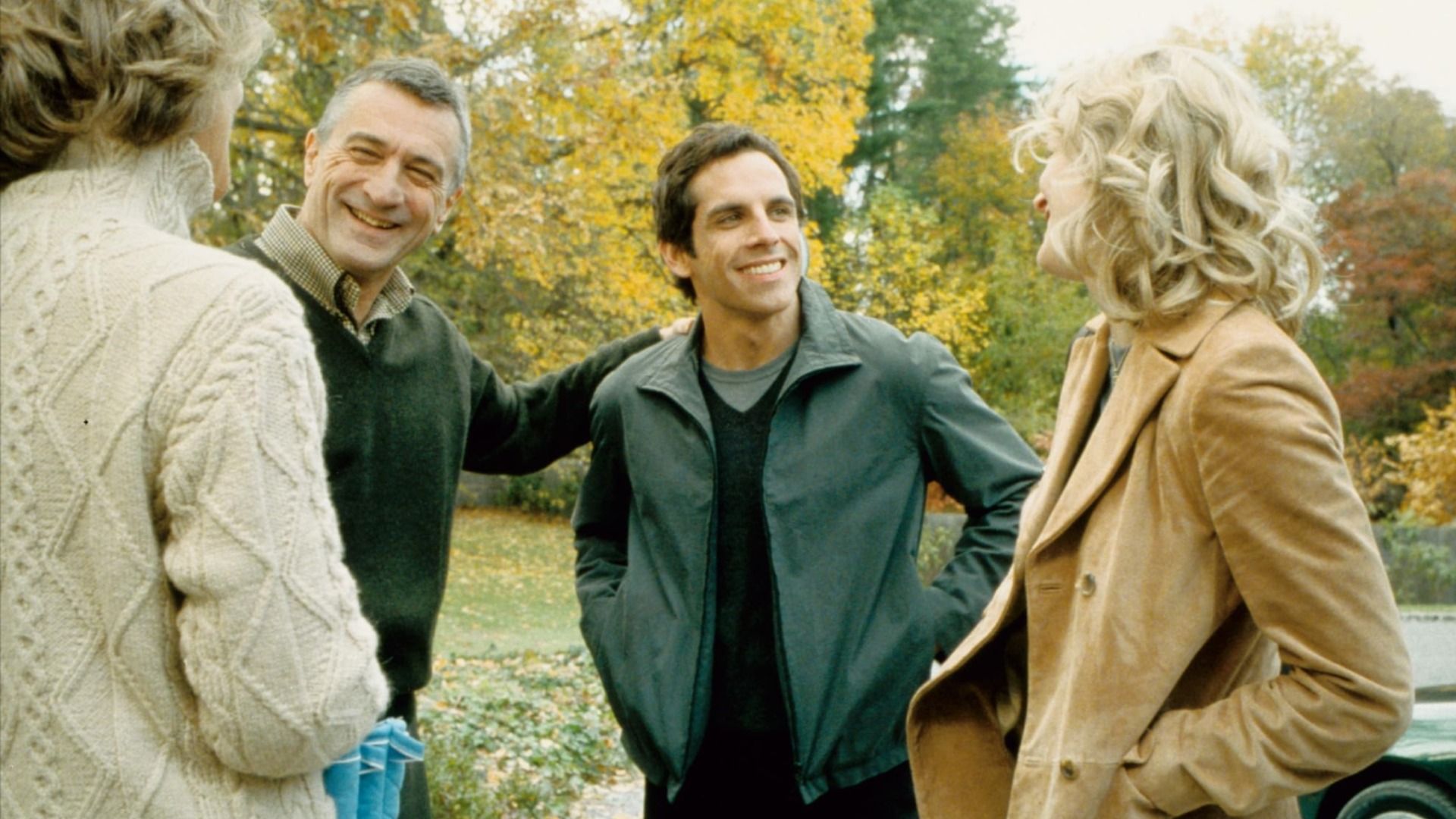 Actorii Robert De Niro într-un pulover verde și Ben Stiller într-o jachetă verde stând lângă copaci cu frunze galbene