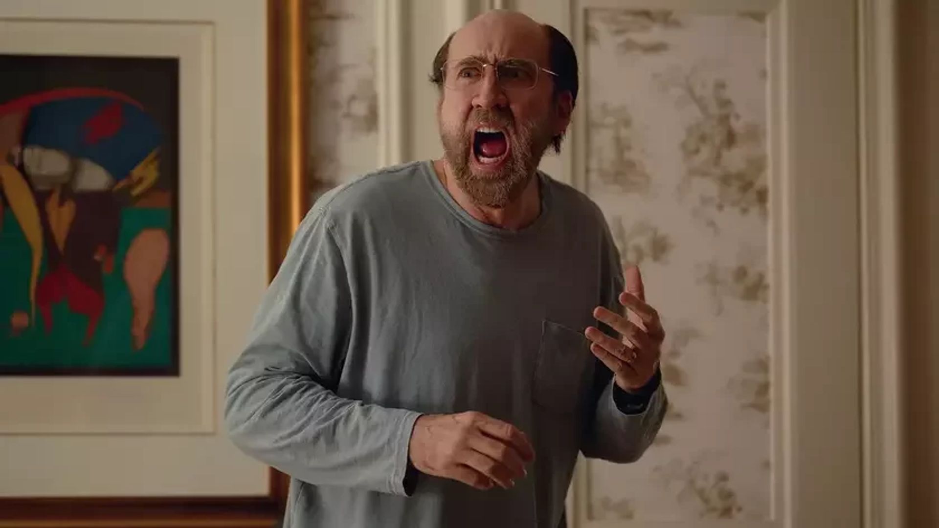 Nicolas Cage purtând ochelari și o cămașă gri cu mâneci lungi țipând în timp ce stă într-o cameră