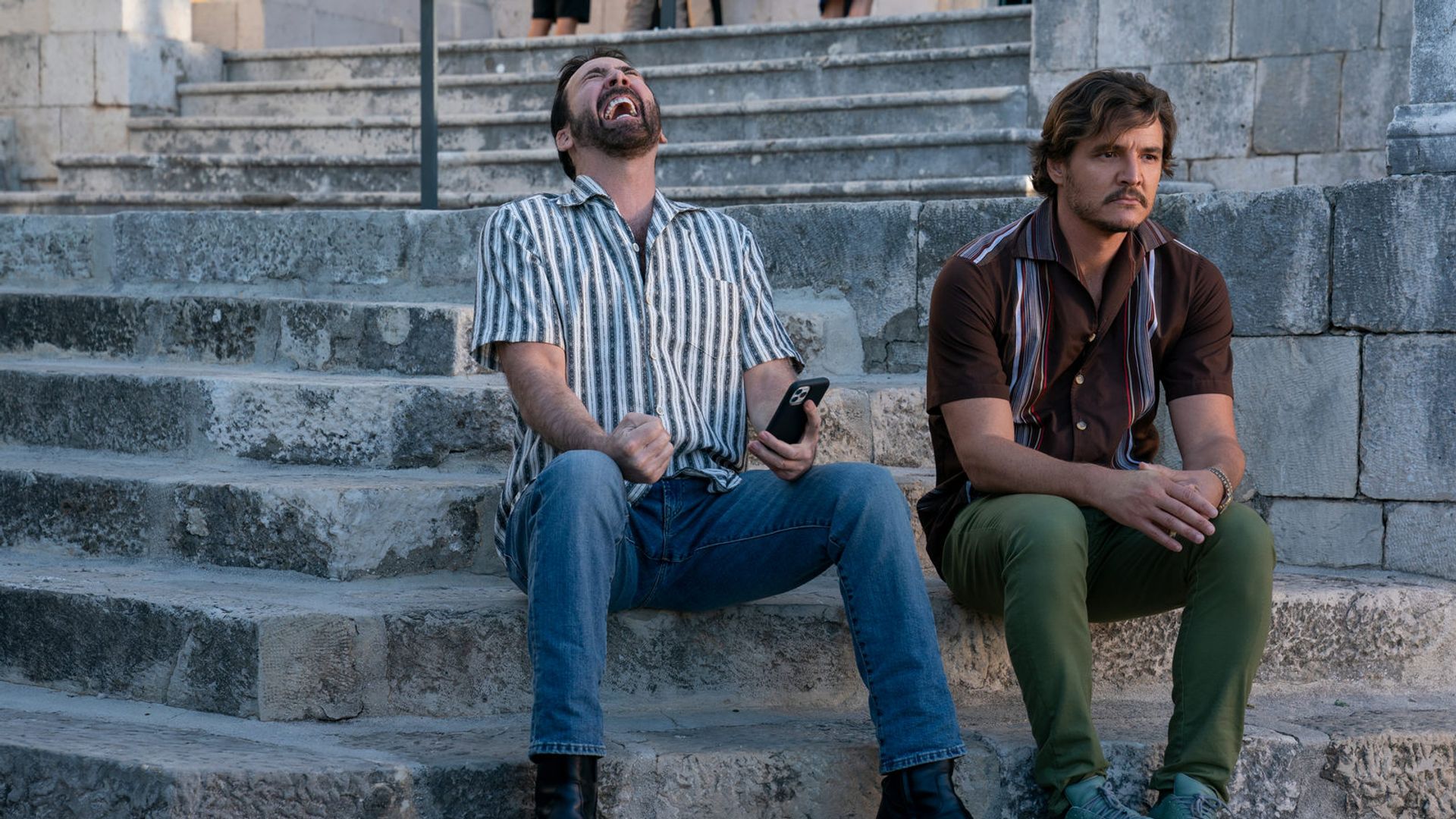 Nicolas Cage și Pedro Pascal stau pe niște trepte de piatră: unul râde cu telefonul în mână, celălalt privește gânditor într-o parte