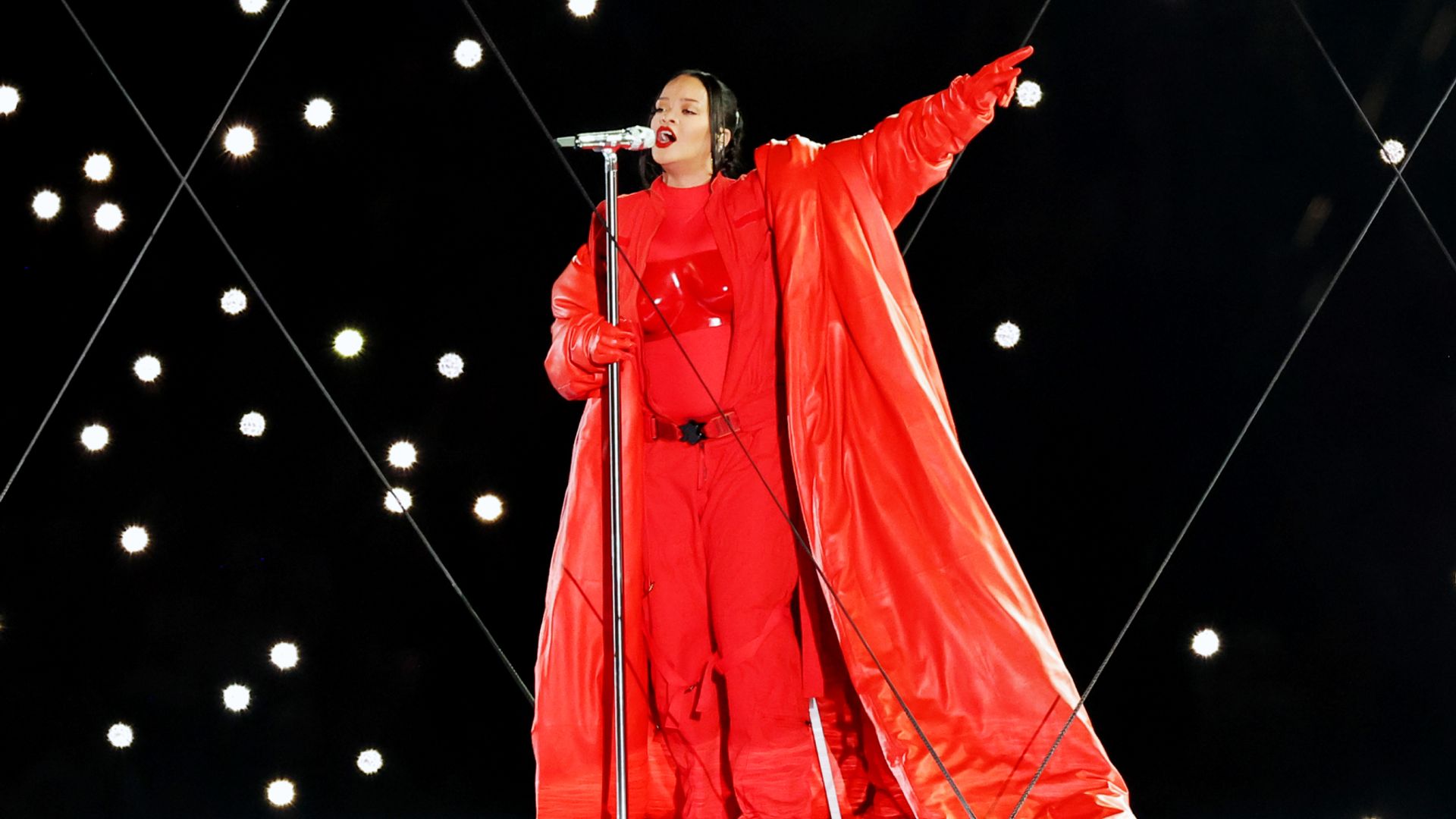 Rihanna într-un costum roșu strălucitor și o pelerină lungă cântă pe scenă cu un microfon