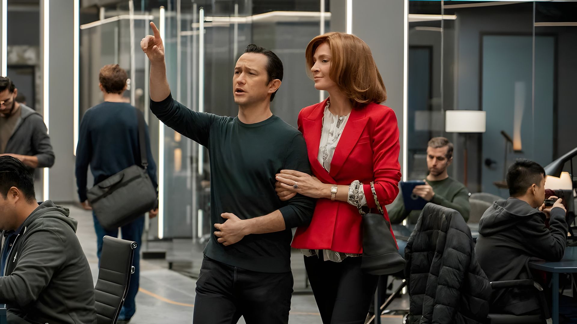 Joseph Gordon-Levitt în haine întunecate arată cu degetul înainte în timp ce Uma Thurman în jachetă roșie se ține de brațul lui