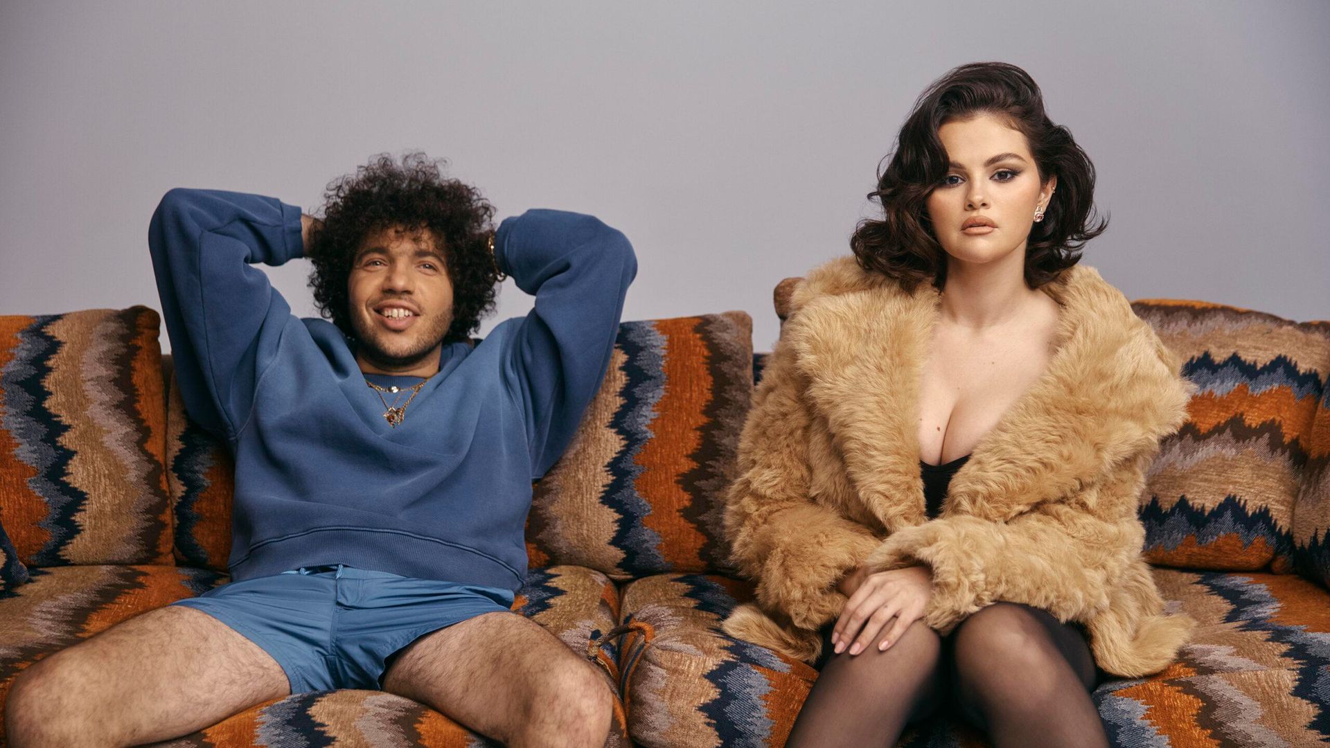 Selena Gomez într-o haină de blană și Benny Blanco într-un hanorac albastru și pantaloni scurți stând relaxați