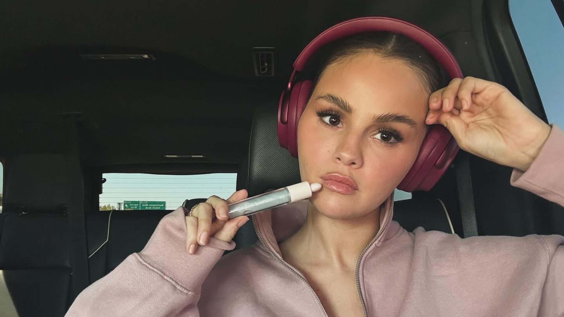 Selena Gomez într-o bluză roz și cu căști pe urechi stă în mașină, ținând un produs cosmetic în fața feței