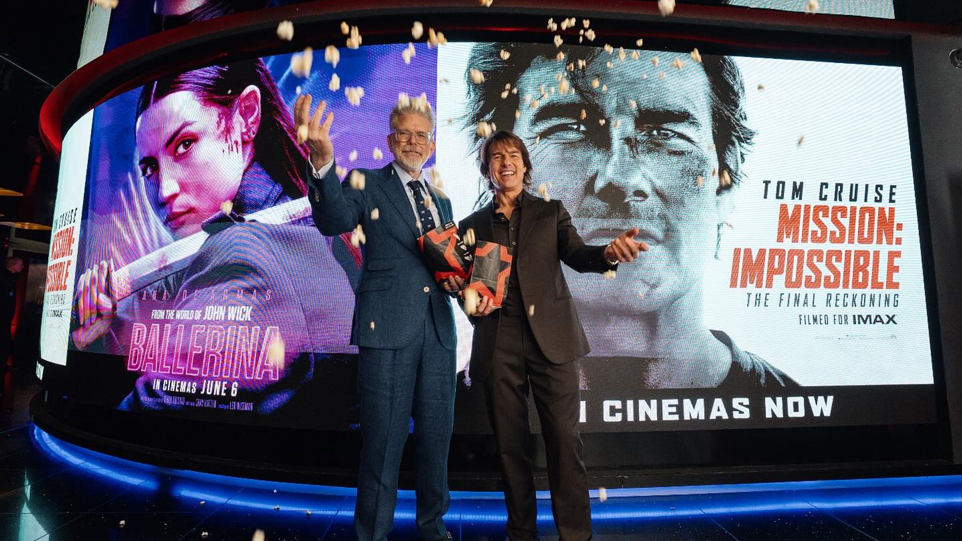 Doi bărbați în costume pozează cu cutii de popcorn pe un fundal de postere pentru filmele Ballerina și Mission: Impossible