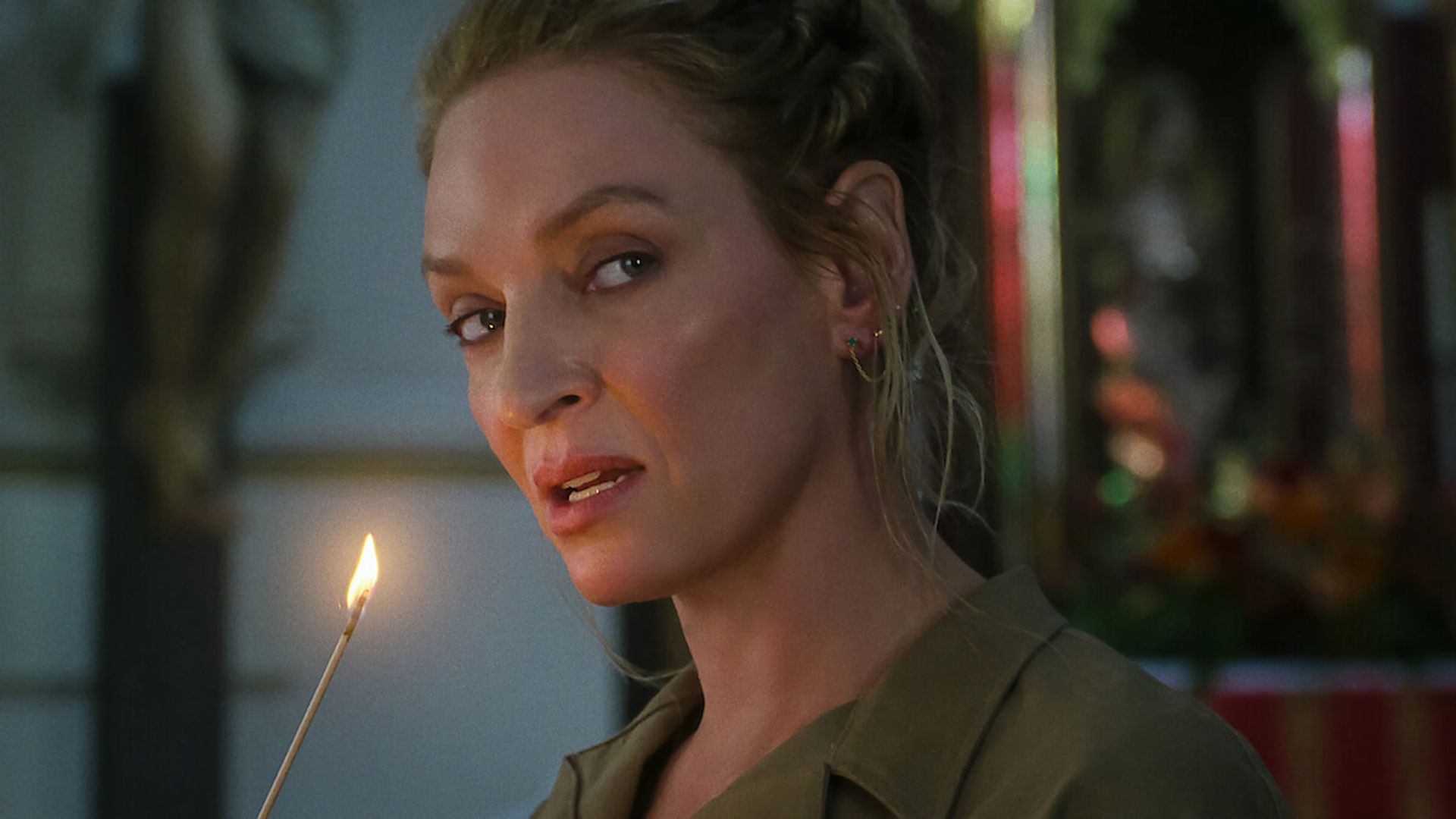Uma Thurman cu părul prins în spate ține o lumânare aprinsă și privește în lateral cu un fundal ușor neclar