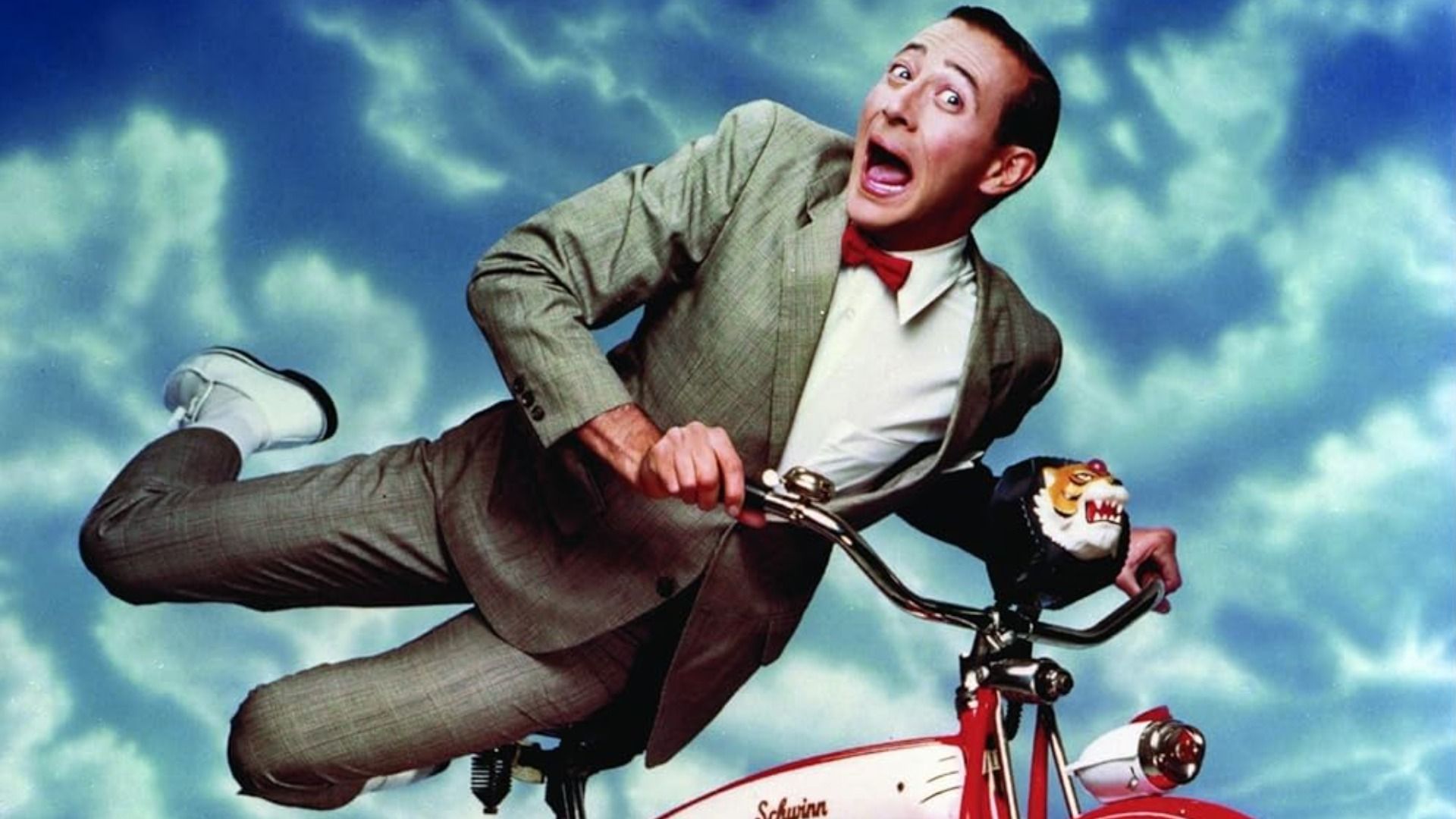 Actorul și comediantul Paul Reubens în costum gri pe o bicicletă