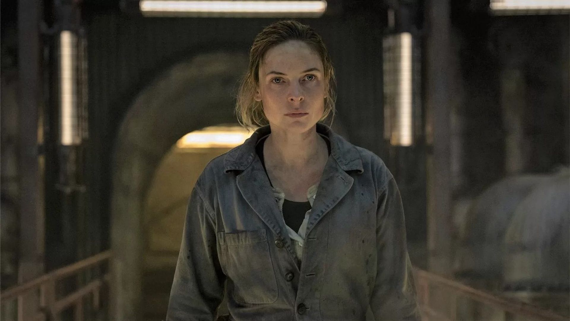 Rebecca Ferguson în salopetă de lucru stă într-un spațiu industrial slab luminat, privind drept înainte