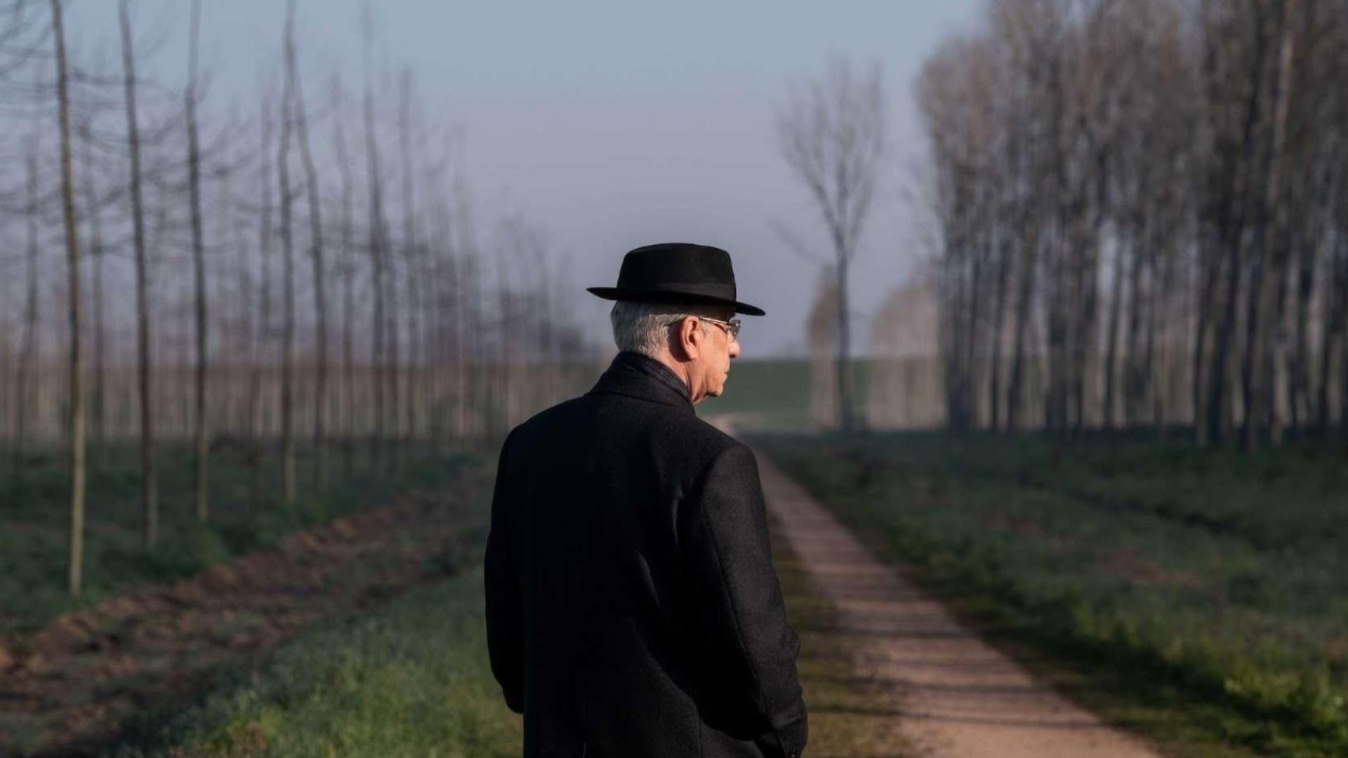 Actorul Toni Servillo într-un palton negru și pălărie neagră stă lângă copaci