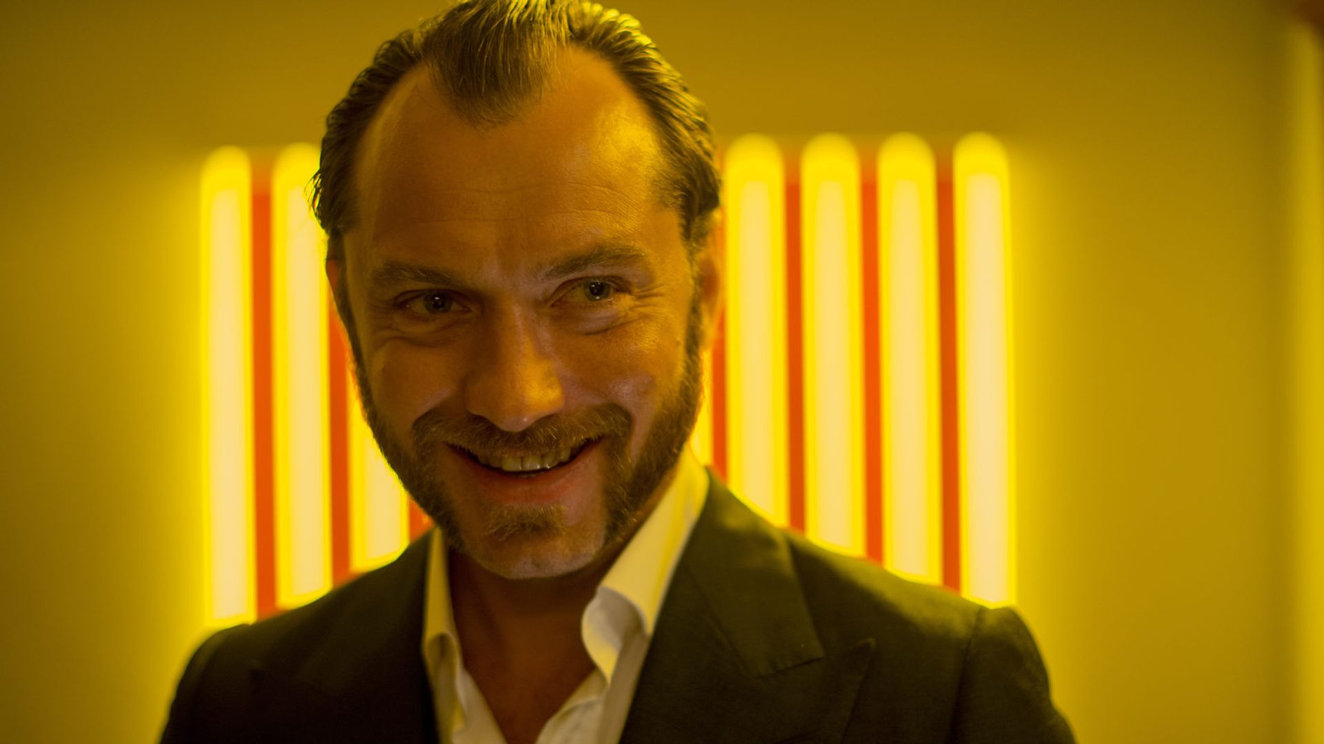 Jude Law într-o jachetă de costum zâmbește pe un fundal cu dungi de lumină galbenă strălucitoare