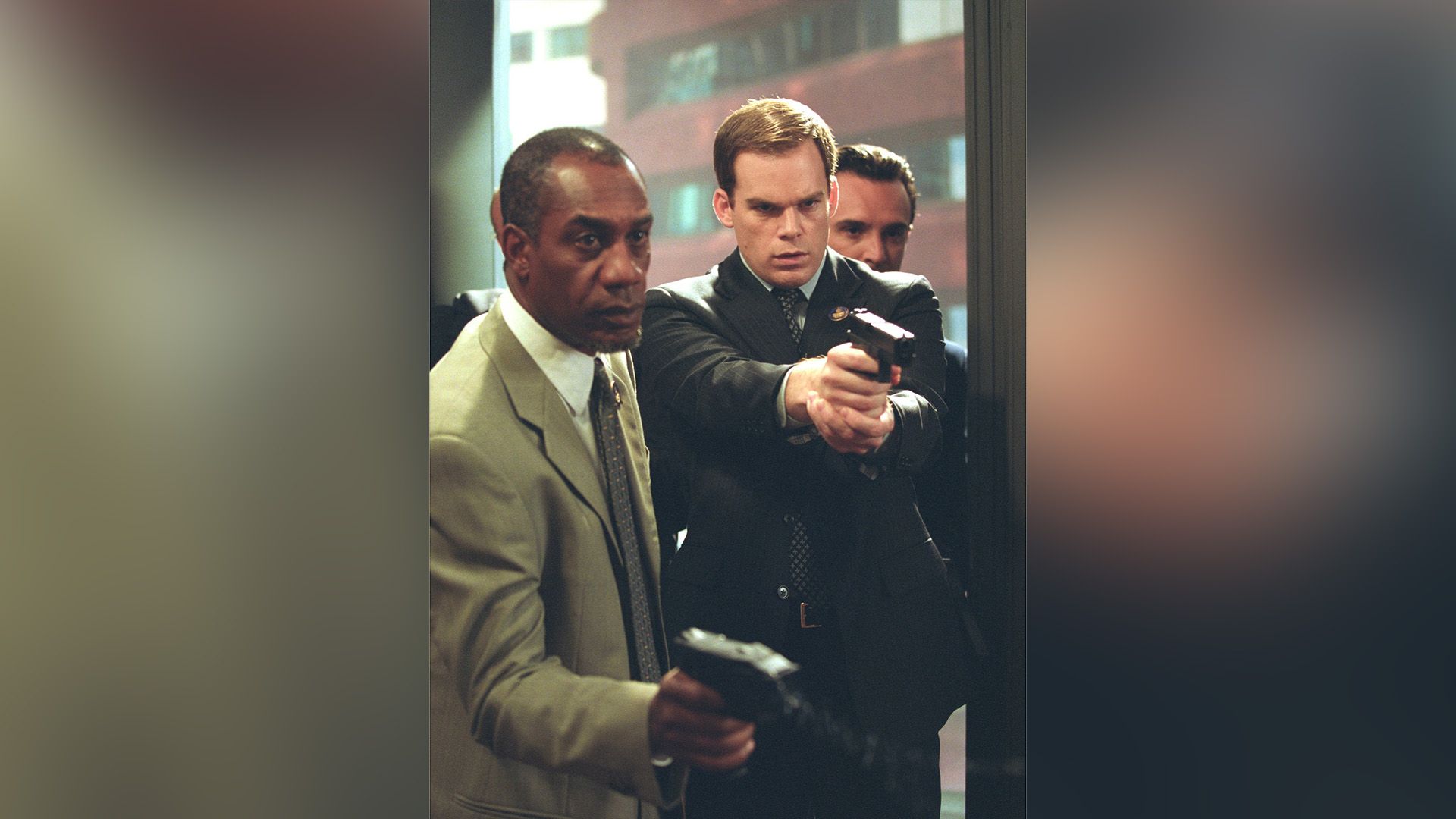 Michael C. Hall și un bărbat de culoare stând cu brațele întinse ținând arme