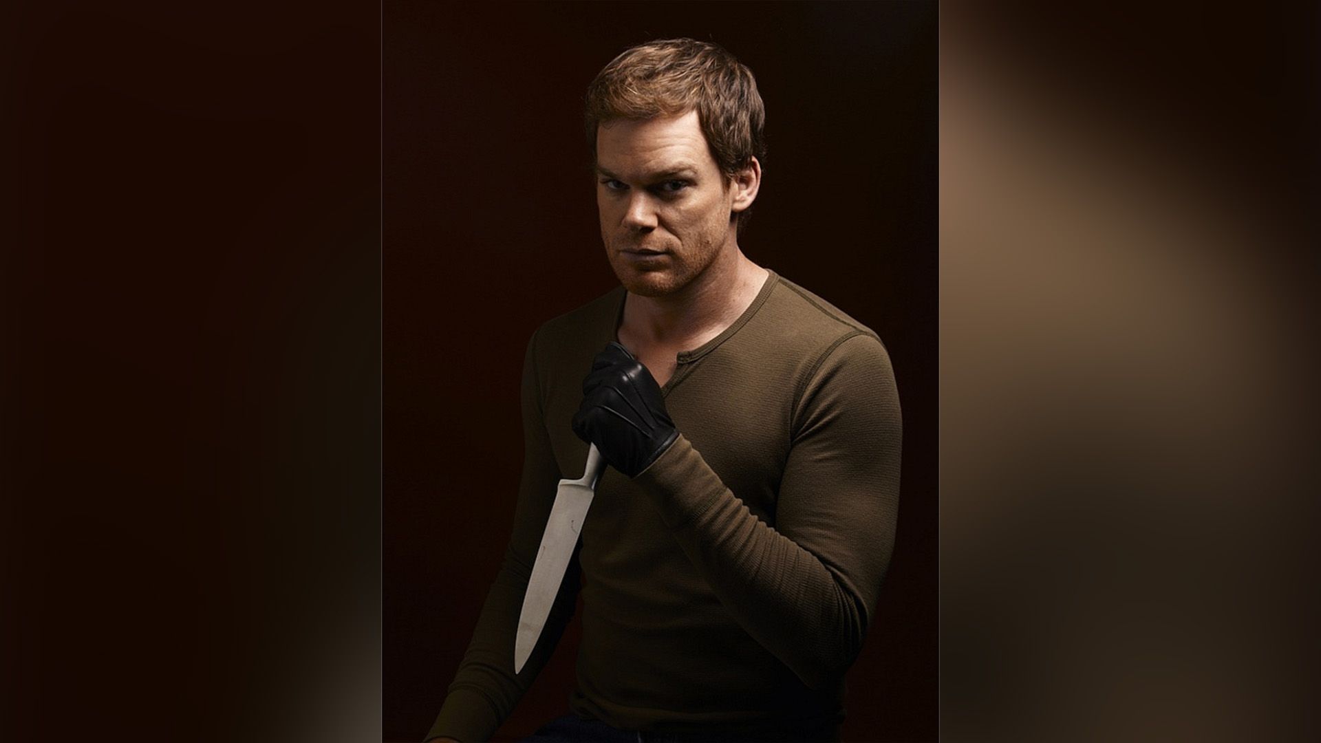 Michael C. Hall într-o jachetă maro și mănuși negre ținând un cuțit în mână