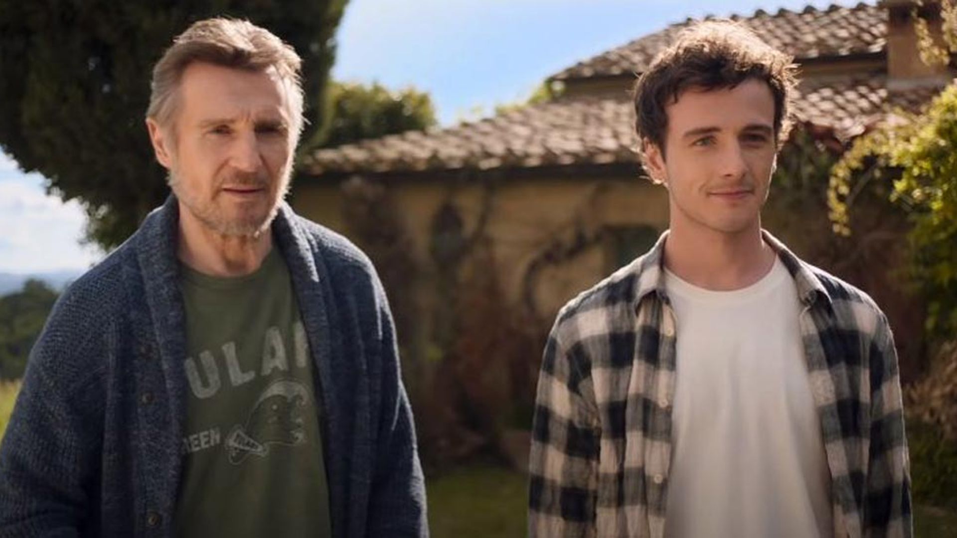 Liam Neeson și Michael Richardson stau unul lângă altul în fața unei case cu acoperiș de țiglă într-o zi însorită