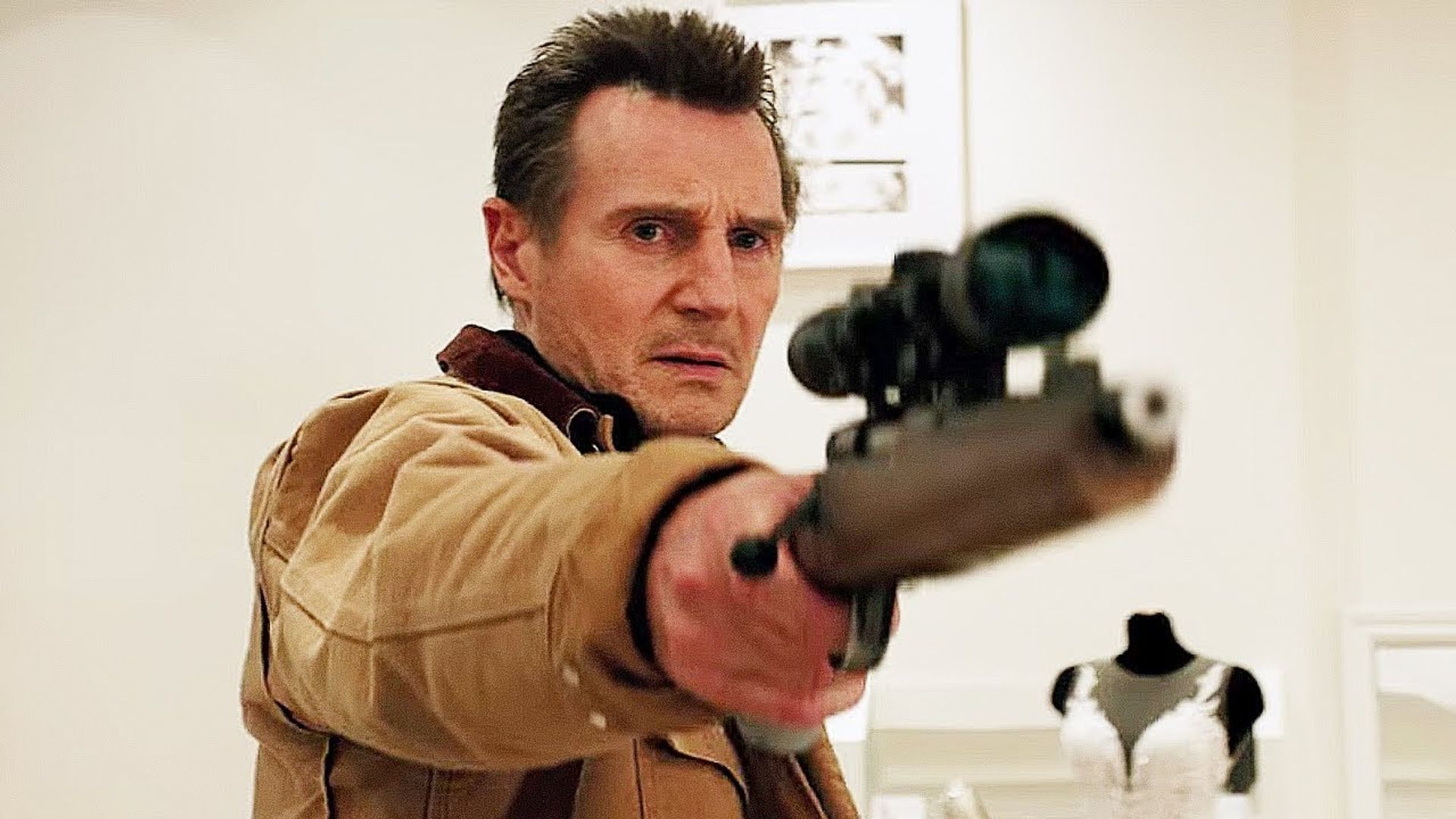 Liam Neeson într-o jachetă bej țintește cu o pușcă cu lunetă, cu un manechin în rochie de mireasă în spatele lui