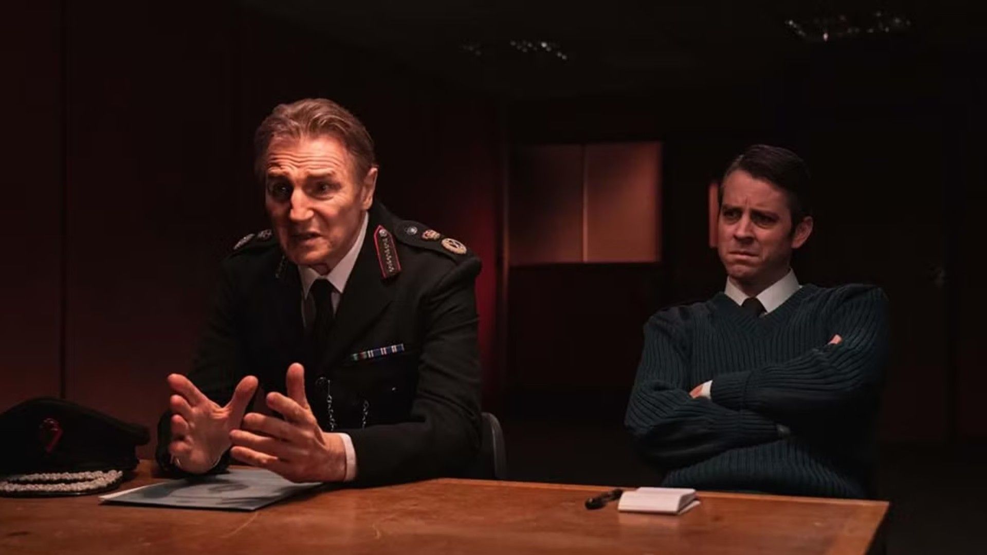 Liam Neeson în uniformă de polițist stând la o masă de interogatoriu, scenă din serialul "Derry Girls"