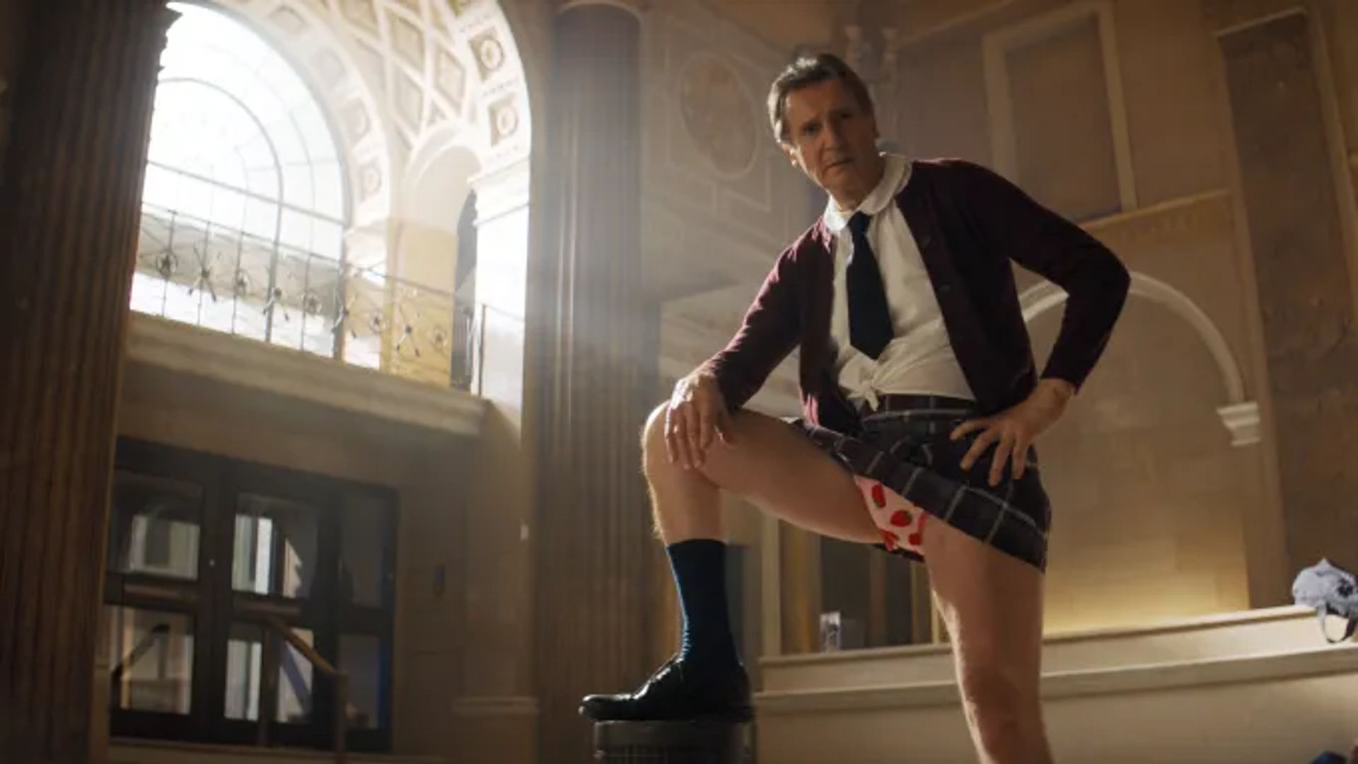 Liam Neeson în uniformă școlară și cravată pozează cu piciorul ridicat, expunând lenjeria intimă cu inimi, pe fondul unei decorațiuni cu coloane
