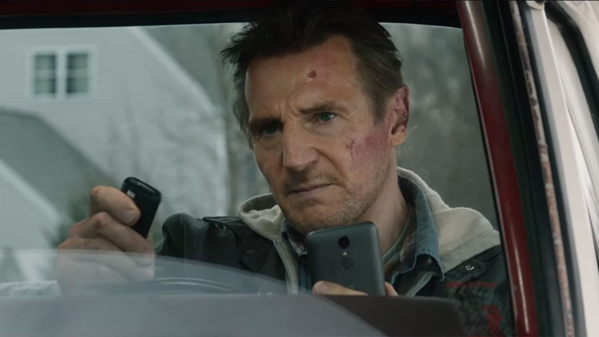 Liam Neeson stă în mașină privind tensionat la telefonul și telecomanda din mâinile sale